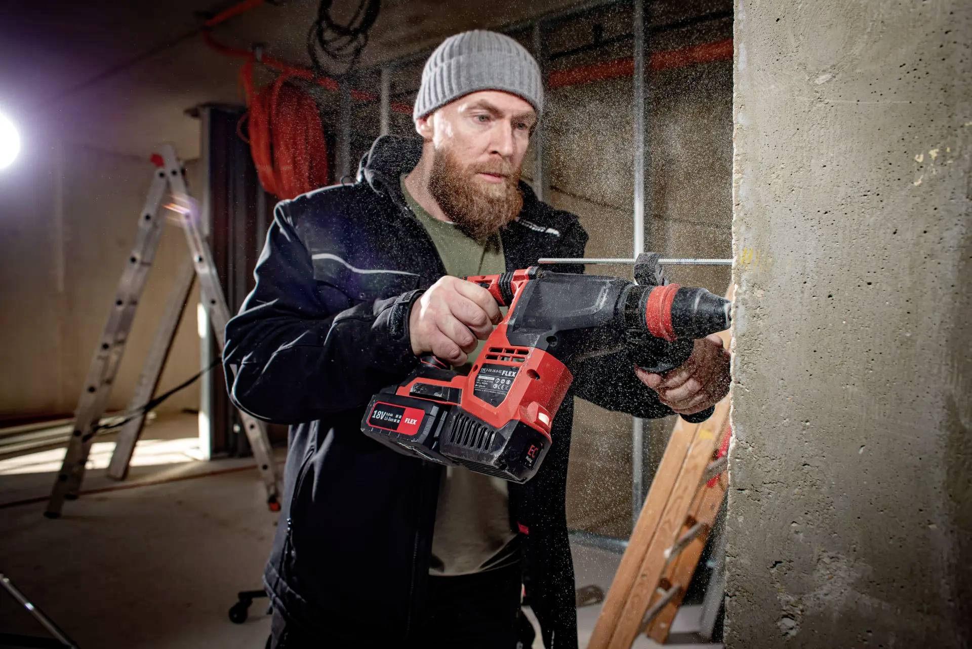 Ein Mann in Arbeitskleidung bohrt mit einem roten Bohrhammer ein Loch in eine Betonwand, hinter ihm ist eine Leiter zu sehen.