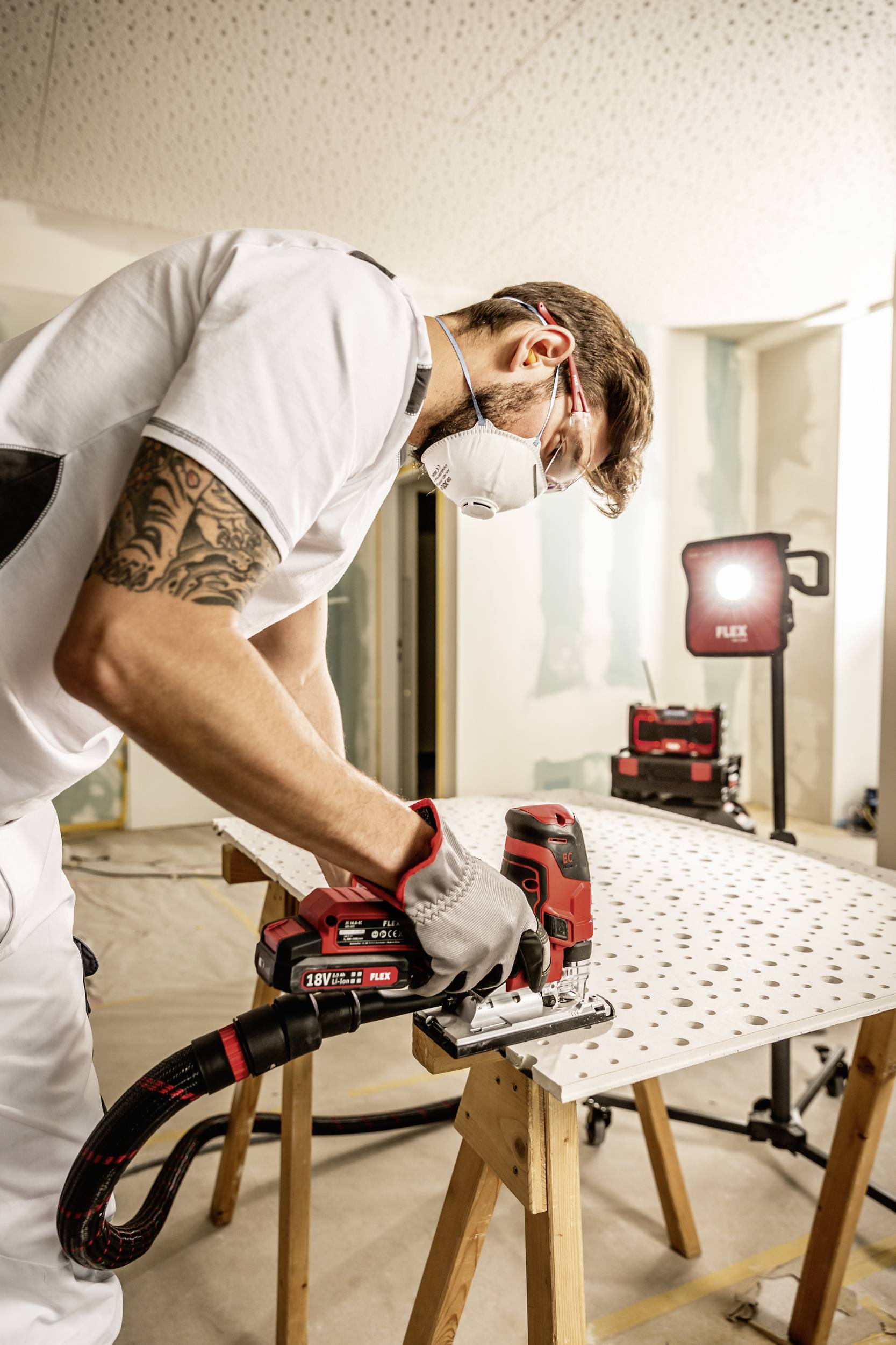 Ein Handwerker sägt mit einer elektrischen Stichsäge ein Holzbrett. Er trägt Arbeitsschutzkleidung, inklusive Maske und Handschuhen.