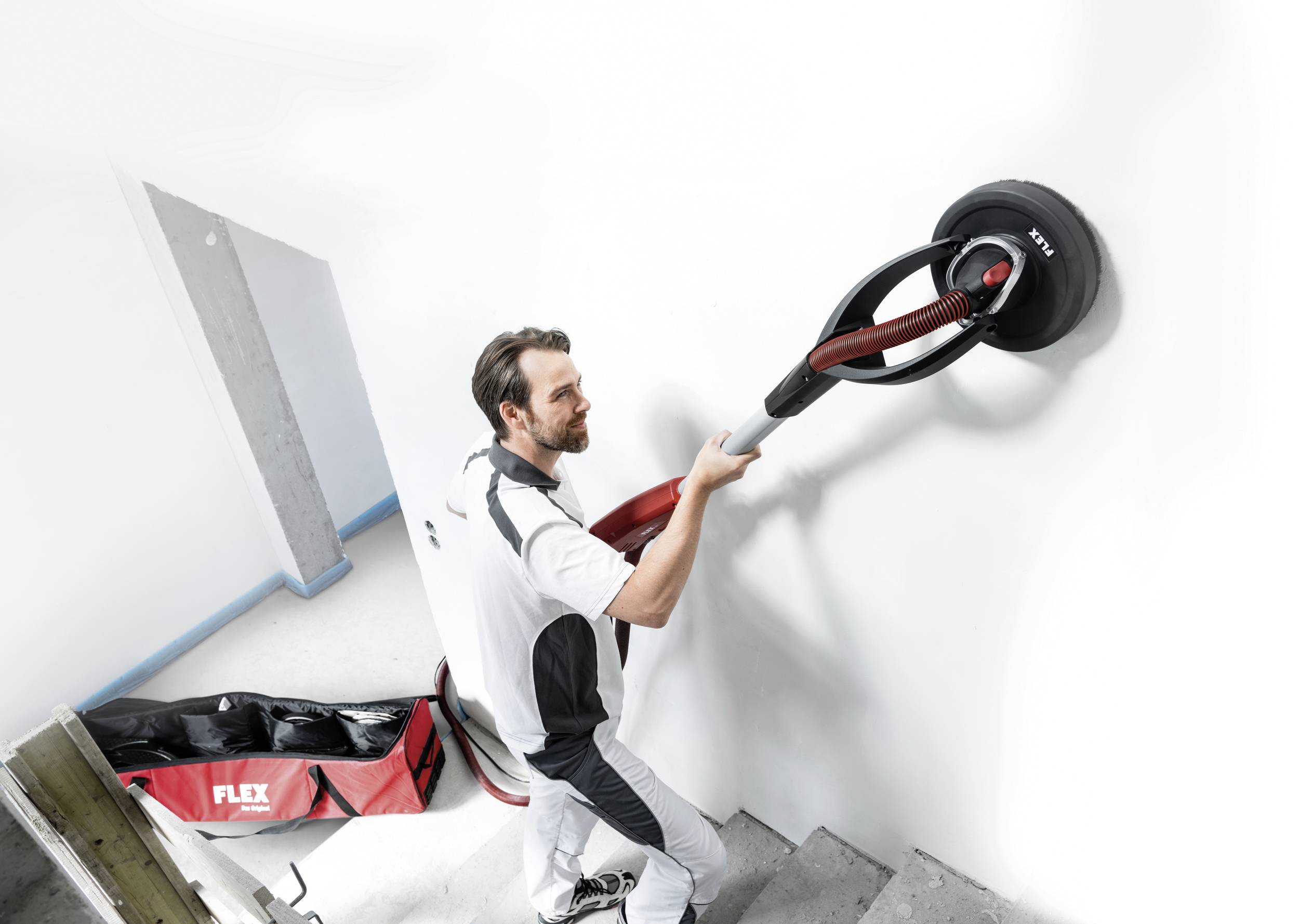 Ein Mann verwendet eine Wandschleifmaschine an einer weißen Wand. Neben ihm liegt eine Tasche mit dem Logo 'FLEX'.