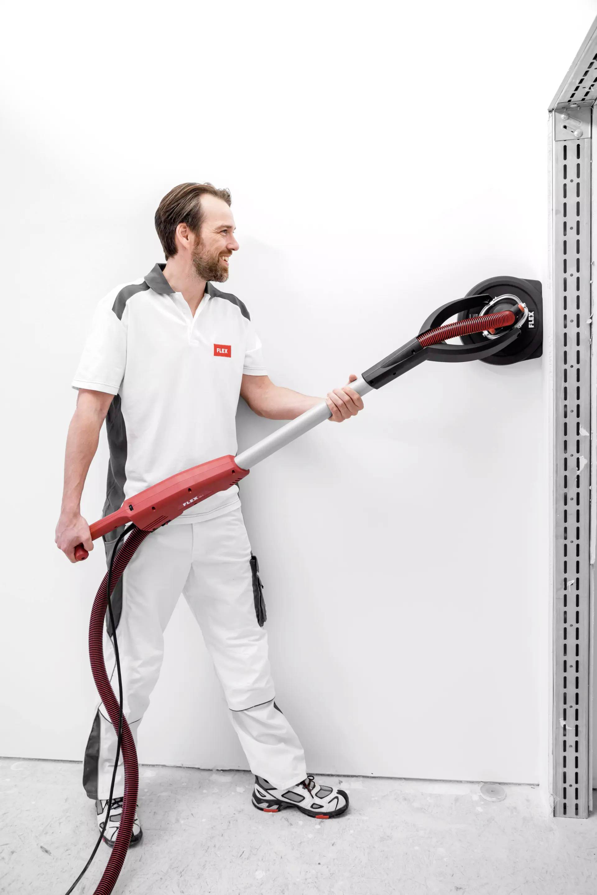 Ein Mann schleift mit einer elektrischen Wand- und Deckenschleifmaschine eine weiße Wand. Er trägt Arbeitskleidung und Sportschuhe.
