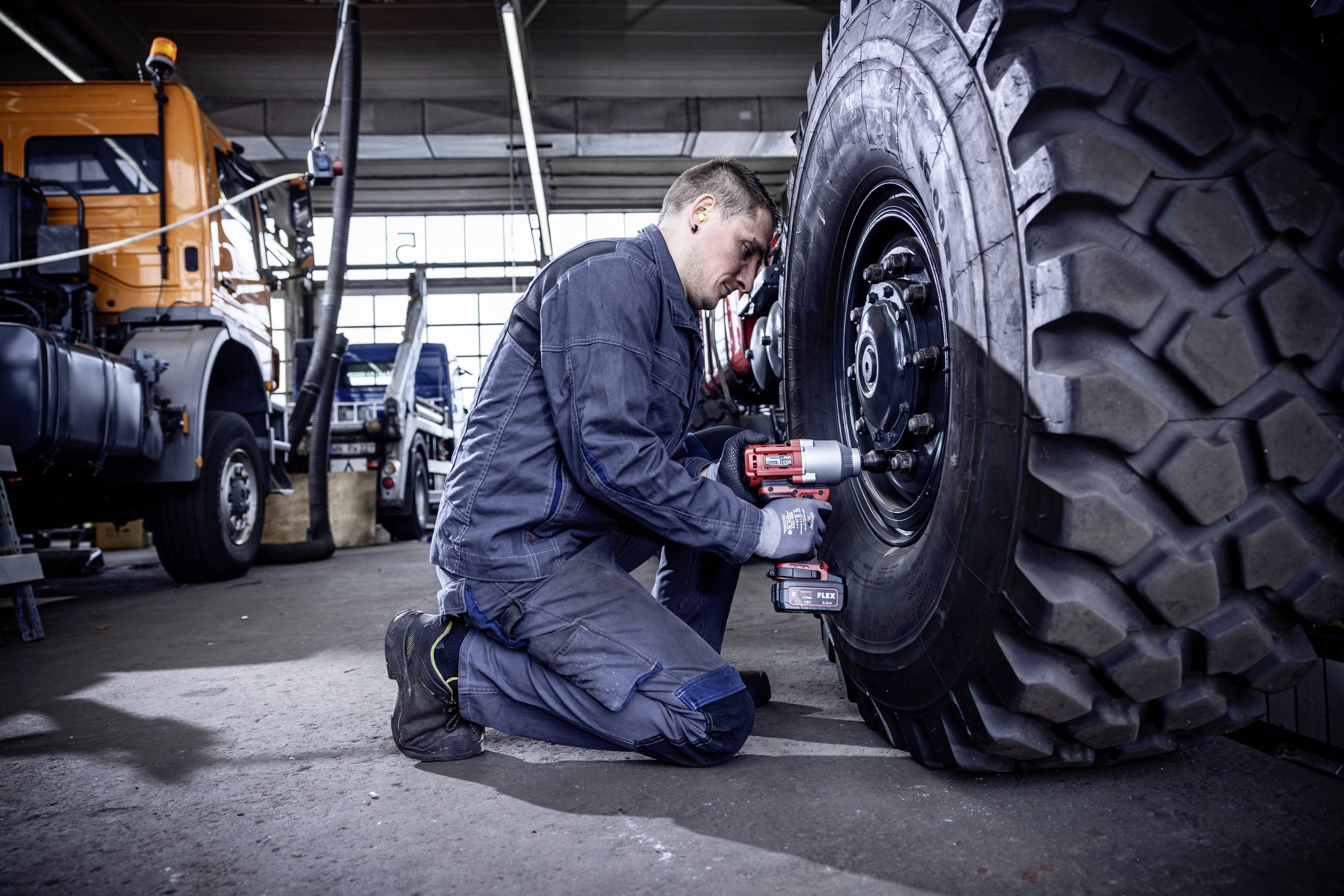 Ein Mechaniker im blauen Overall kniet in einer Werkstatt und repariert mit einem Elektrowerkzeug ein großes Reifenrad eines Lastwagens.