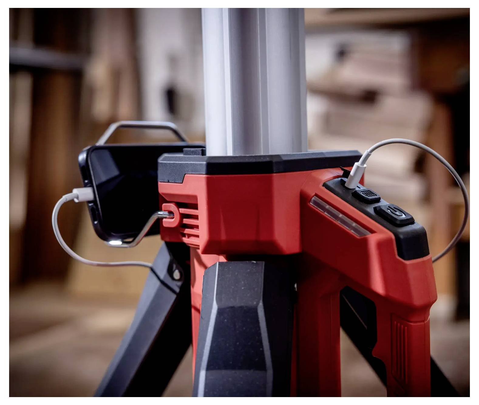 Nahaufnahme eines roten Stativs mit schwarzem Smartphone, das an der Seite befestigt ist. Szenerie zeigt eine Werkstattumgebung.