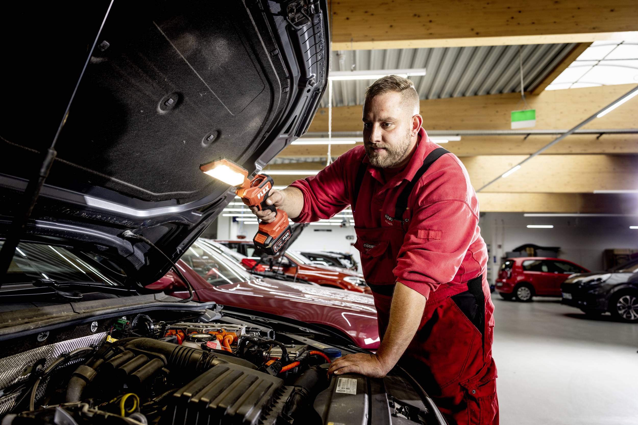 Ein Automechaniker in roter Arbeitskleidung inspiziert mit einer Lampe den Motor eines Fahrzeugs in einer Werkstatt.