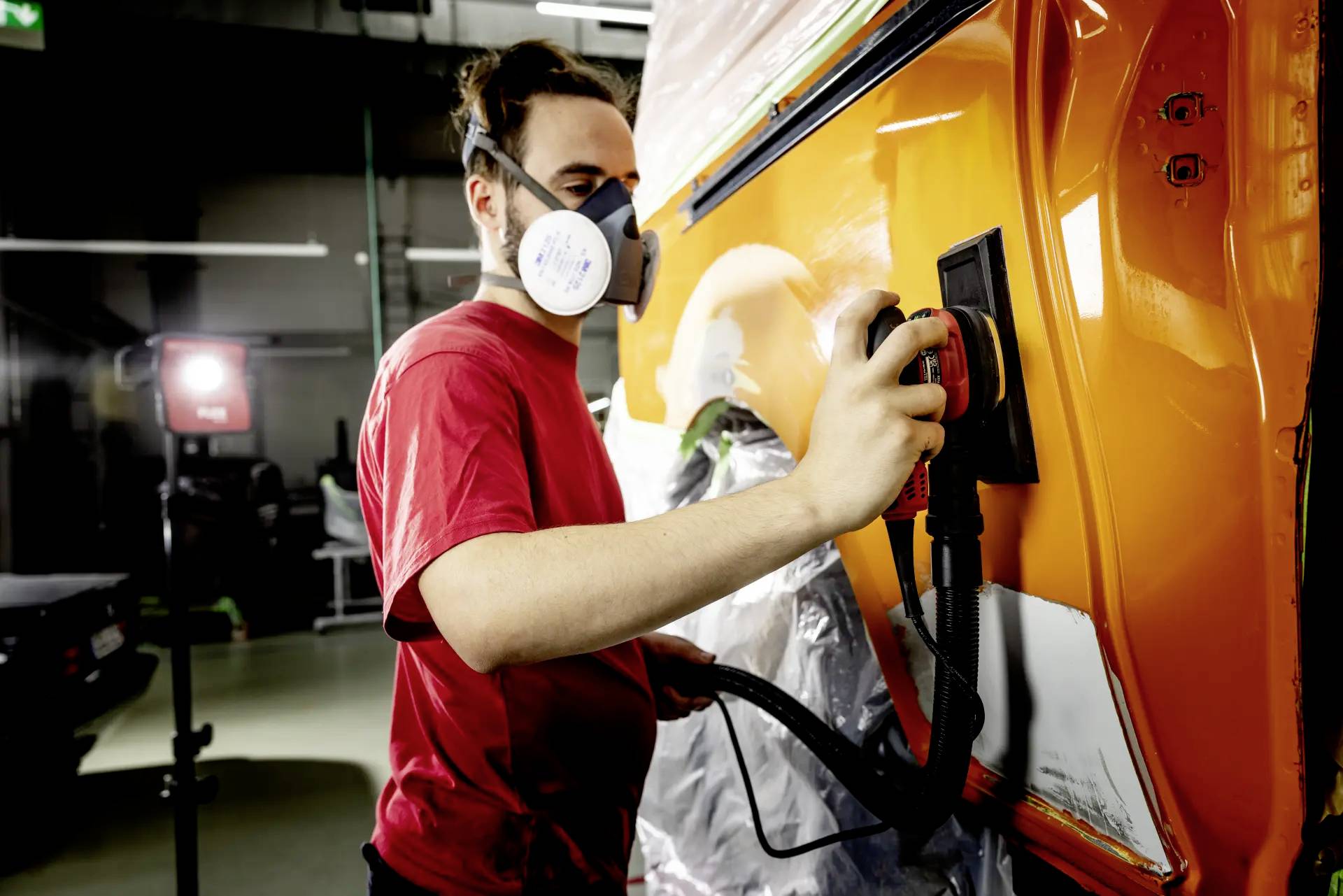 Eine Person trägt eine Atemschutzmaske und schleift die orangefarbene Heckklappe eines Autos in einer Werkstatt.