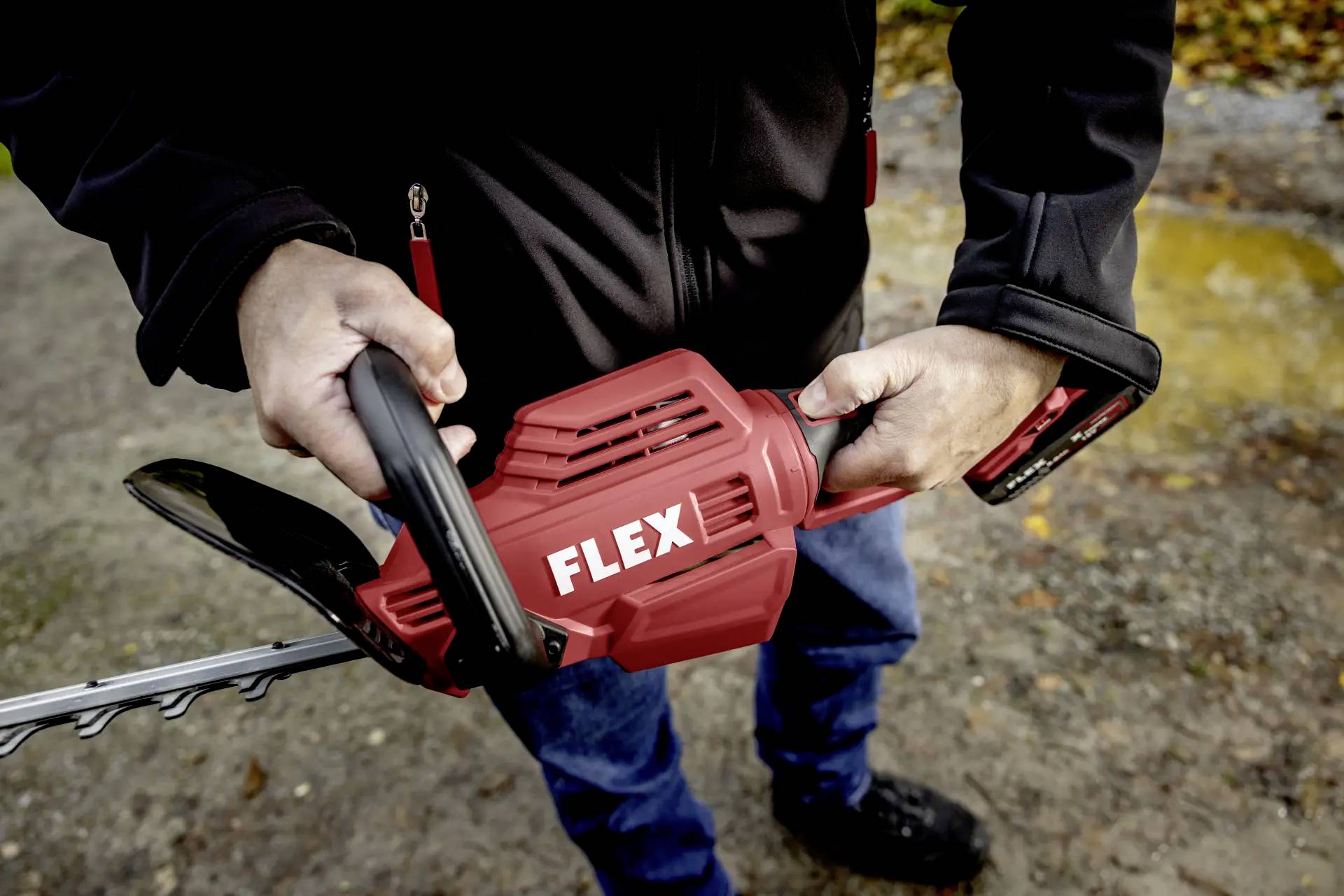 Eine Person hält eine rote, elektrische Heckenschere mit der Aufschrift 'FLEX'. Sie steht draußen auf einem sandigen Boden.