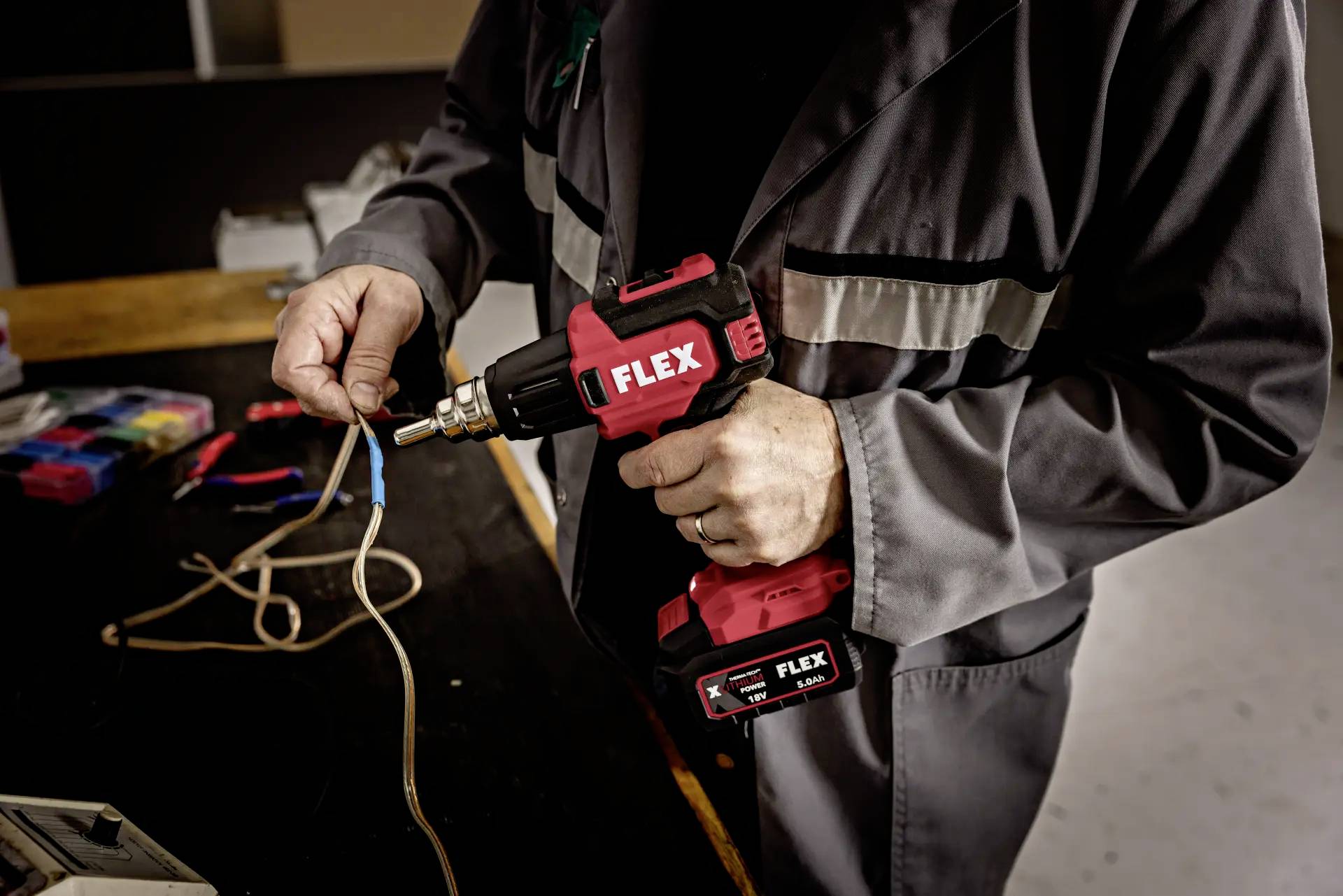 Person in Arbeitskleidung benutzt eine rote Bohrmaschine, um ein Kabel an einem Tisch zu bohren oder zu bearbeiten.