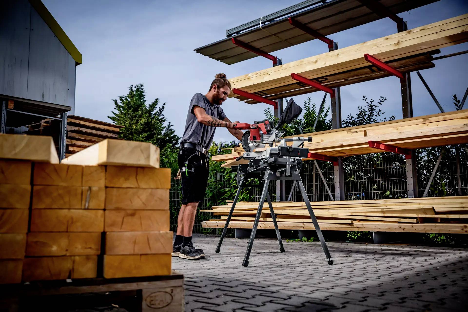 Eine Person arbeitet draußen mit einer Gehrungssäge, umgeben von gestapeltem Holz und Regalen mit Balken, in einem Holzlager.
