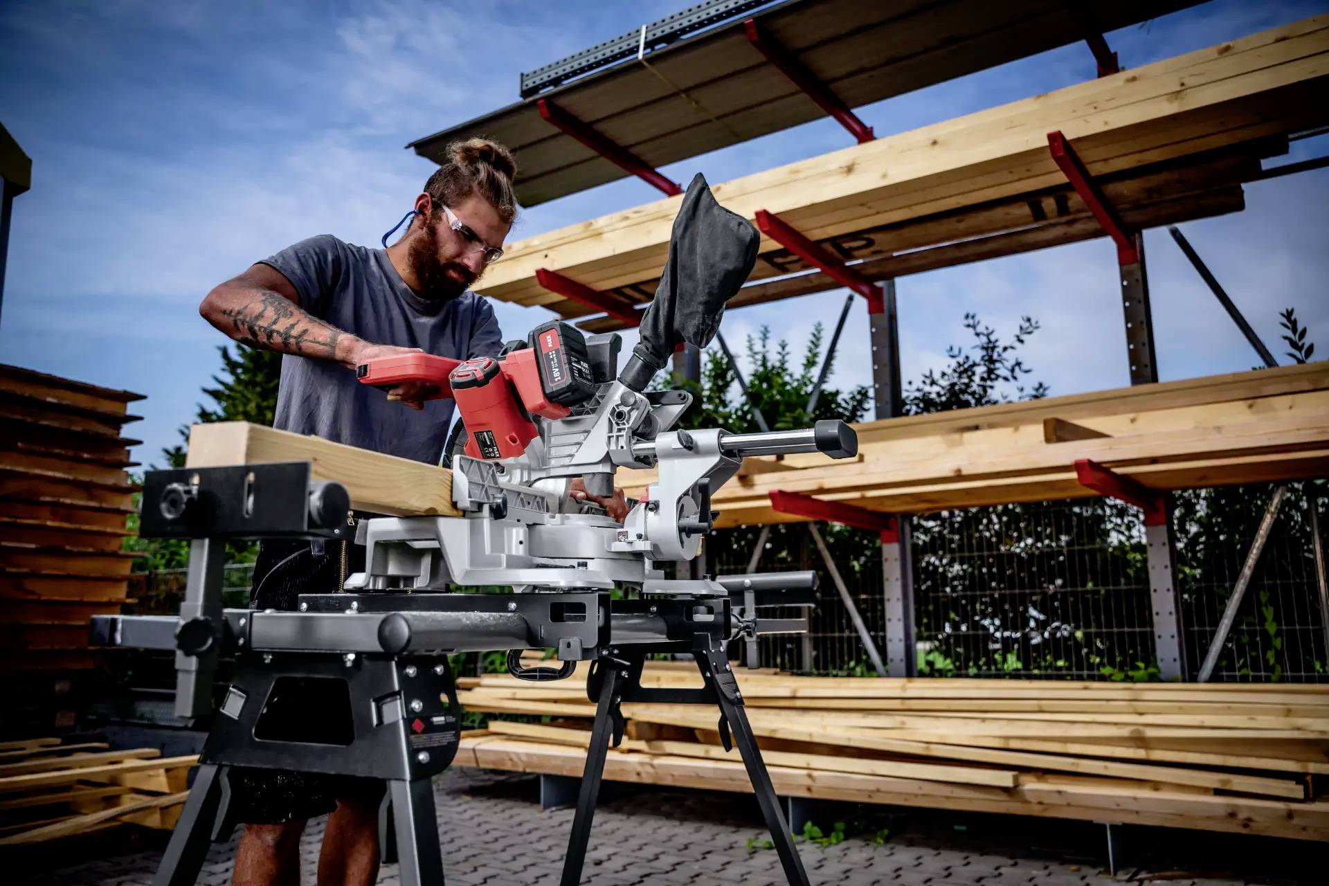 Arbeiter nutzt eine elektrische Säge, um Holz zu schneiden. Im Hintergrund Holzregale im Freien. Klare Vorbereitungen auf Baustelle.