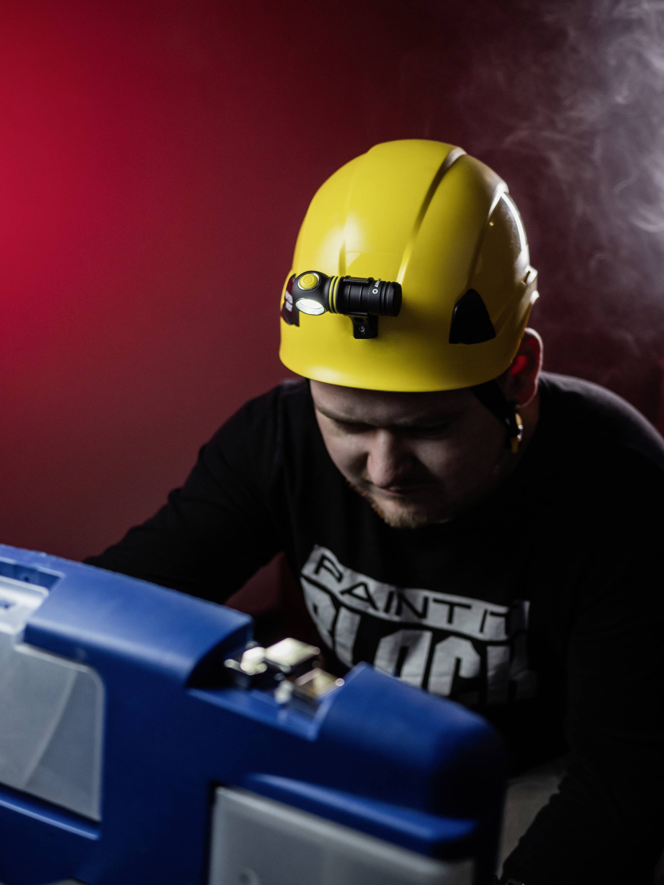 Ein Mann mit gelbem Schutzhelm und Stirnlampe arbeitet konzentriert an einem blauen Gerät vor einem roten Hintergrund.