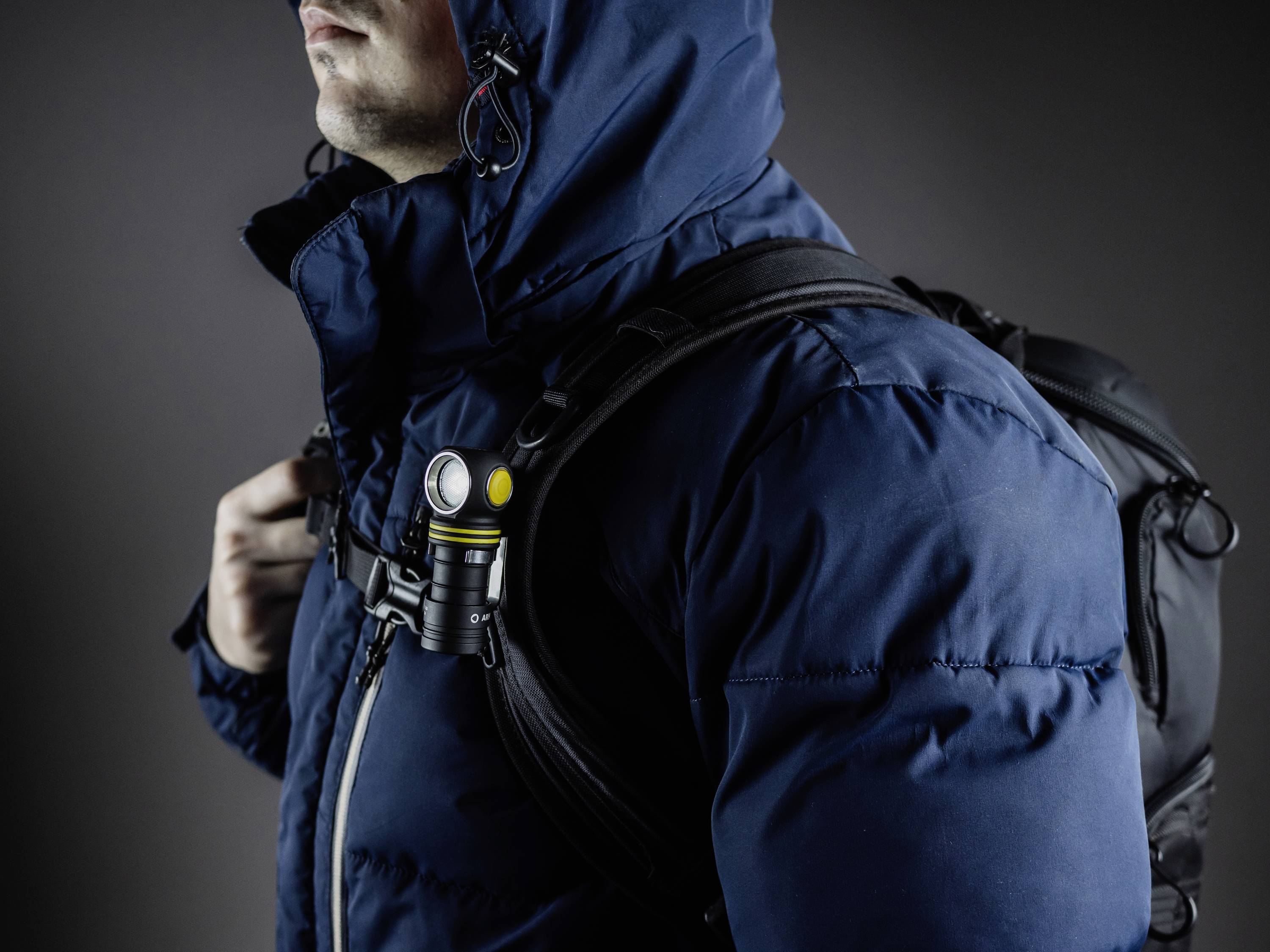 Person in blauer Jacke und Rucksack, trägt eine Taschenlampe am Schulterriemen.