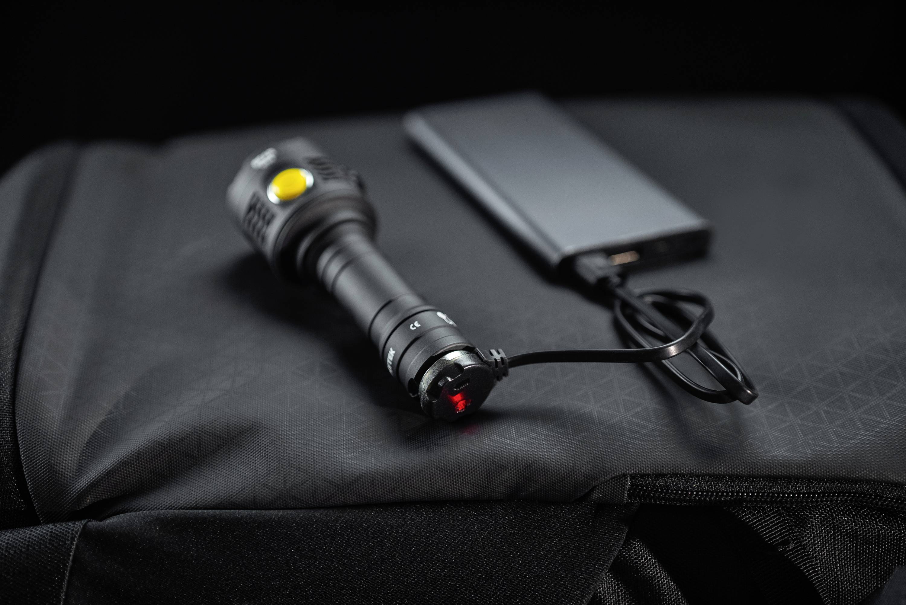 Eine Taschenlampe wird über ein USB-Kabel mit einer Powerbank geladen, alles liegt auf einer schwarzen Tasche.