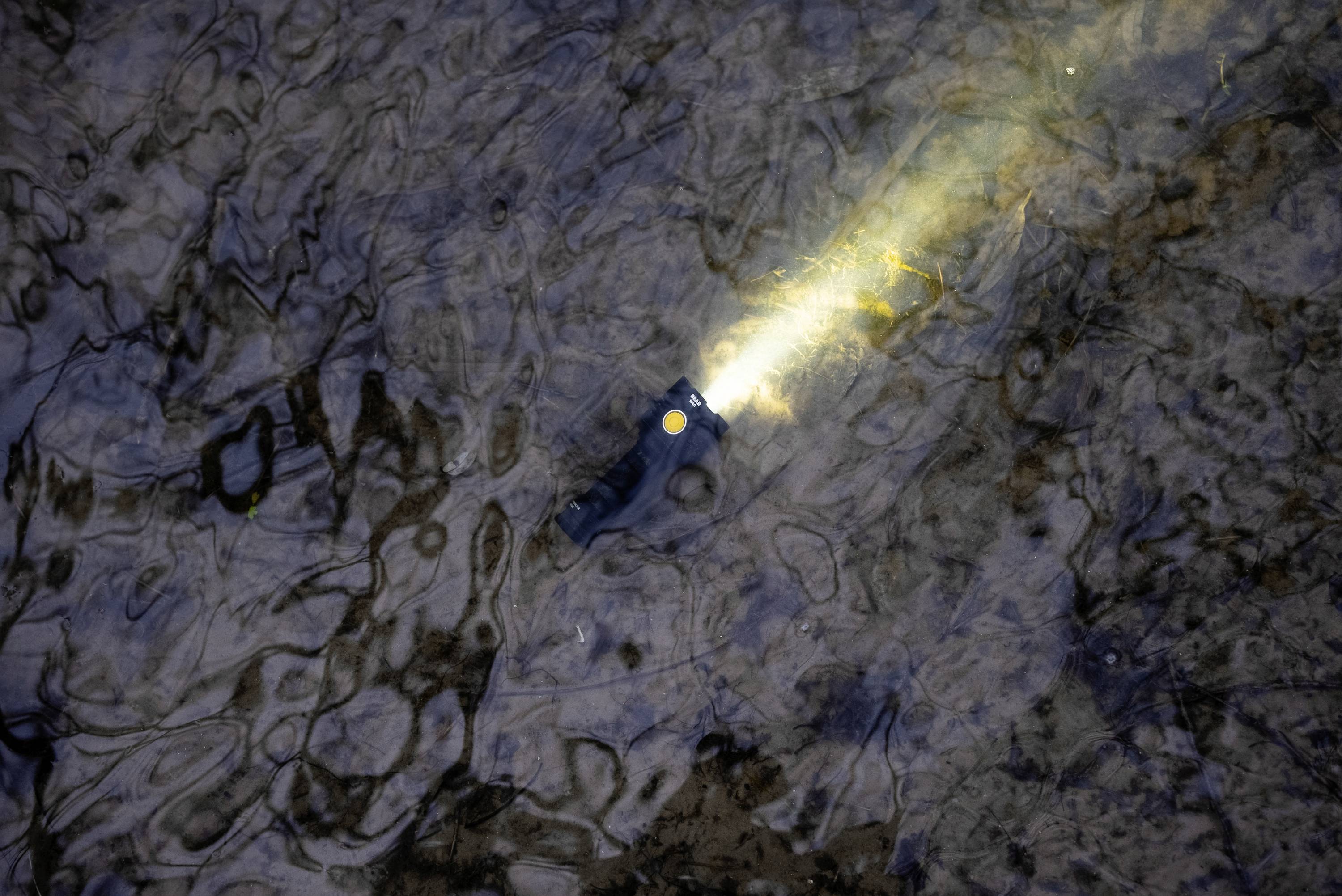 Eine Taschenlampe leuchtet unter Wasser und wirft einen Lichtstrahl nach rechts oben. Sie liegt auf einem steinigen, flachen Flussbett.