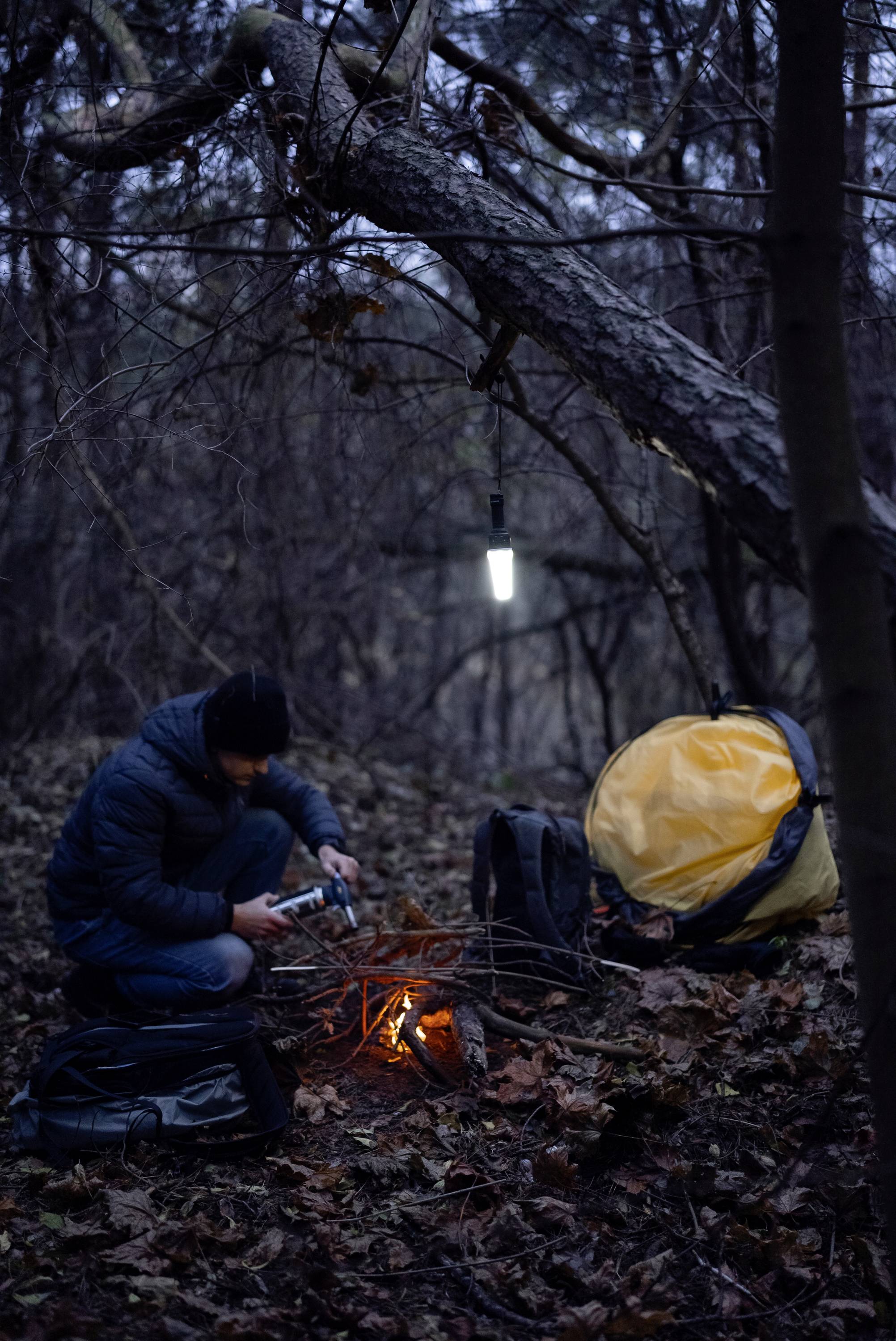 'Person zündet Lagerfeuer im Wald an, mit Zelt und Taschen in der Nähe, bei Dämmerung.'
