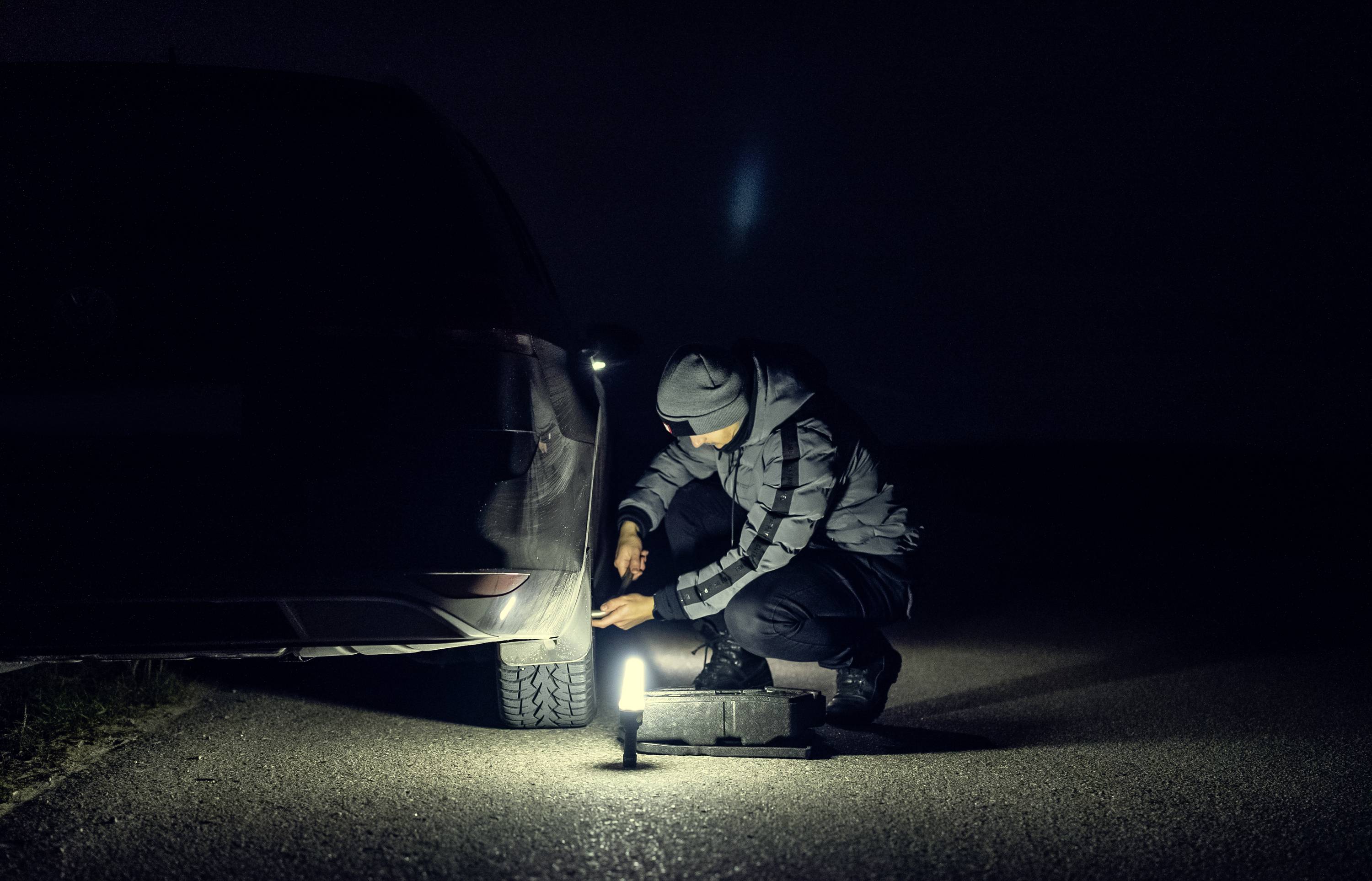 Eine Person wechselt nachts am Straßenrand ein Reifen eines Autos mit einer Laterne als Lichtquelle.