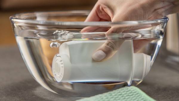 Eine Hand taucht eine Pillendose in eine Schüssel Wasser. Eine grüne Stofffläche liegt im Vordergrund auf der grauen Oberfläche.