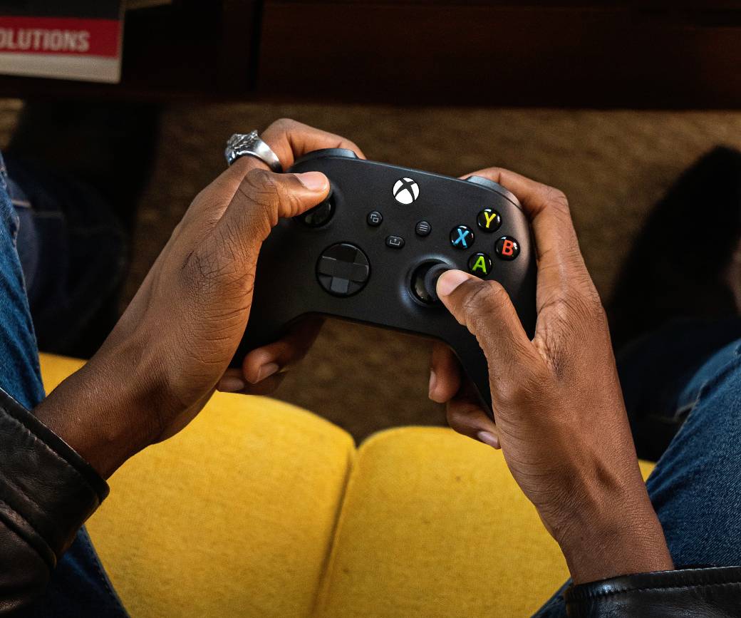Eine Person hält einen schwarzen Videospiel-Controller mit farbigen Tasten auf einem gelben Sofa sitzend, bereit zu spielen.