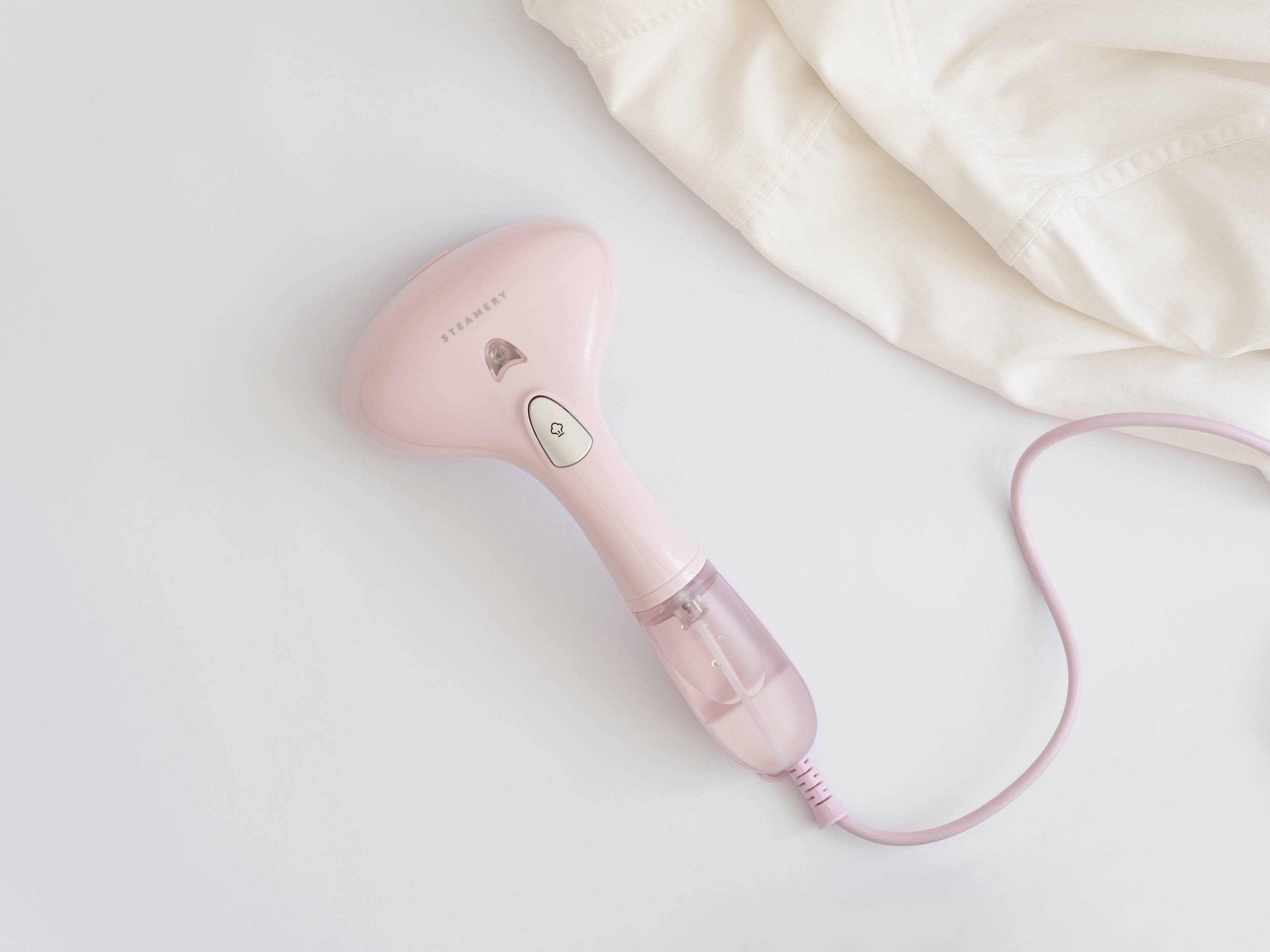 Ein rosa Handdampfer neben einem weißen Hemd auf einer hellen Oberfläche. Der Dampfer ist eingesteckt und einsatzbereit.