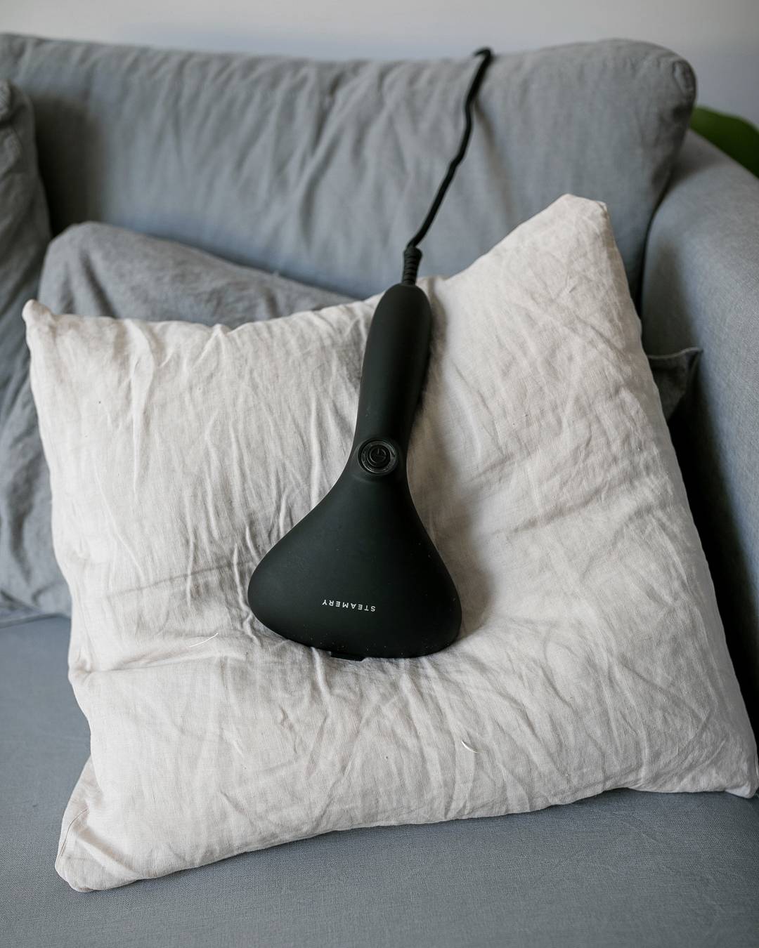Ein schwarzer Handdampfer liegt auf einem beigen Kissen auf einem grauen Sofa.