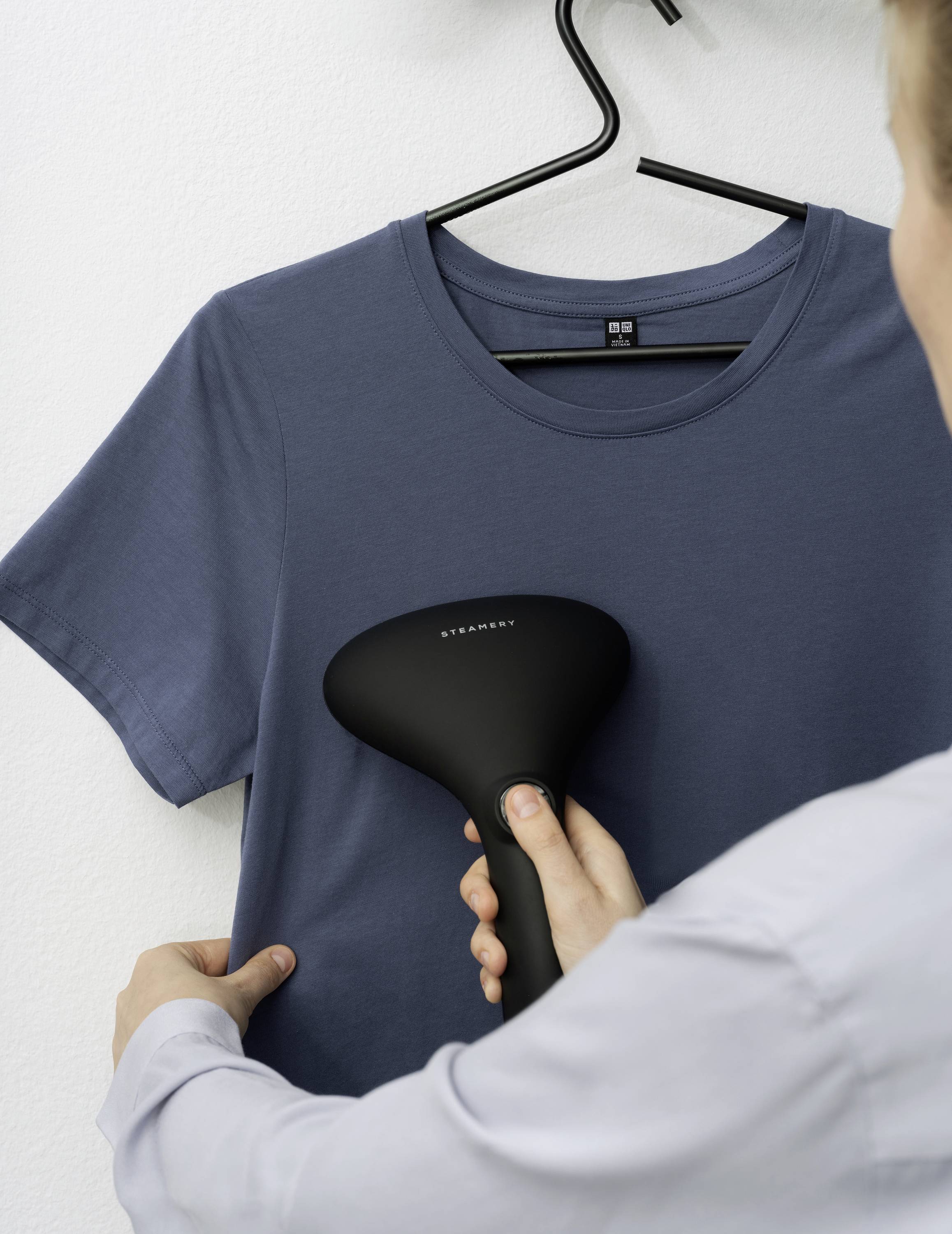 Eine Person bedampft ein blaues T-Shirt mit einem Hand-Dampfglätter, das auf einem Kleiderbügel hängt.