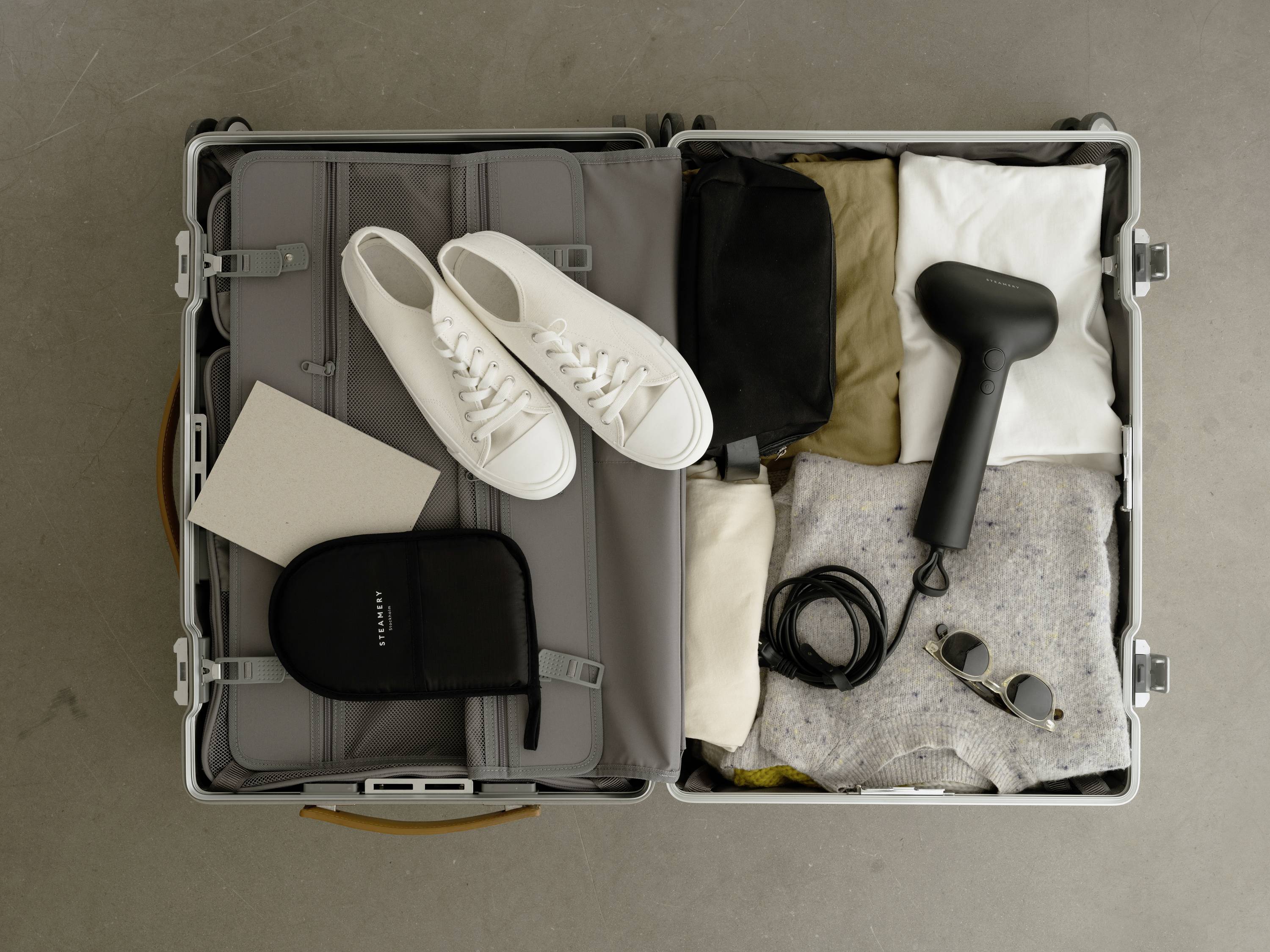 Ein geöffneter Koffer zeigt Kleidung, Sneaker, eine Haarbürste, eine Tasche, Sonnenbrille und Kabel, die ordentlich verstaut sind.