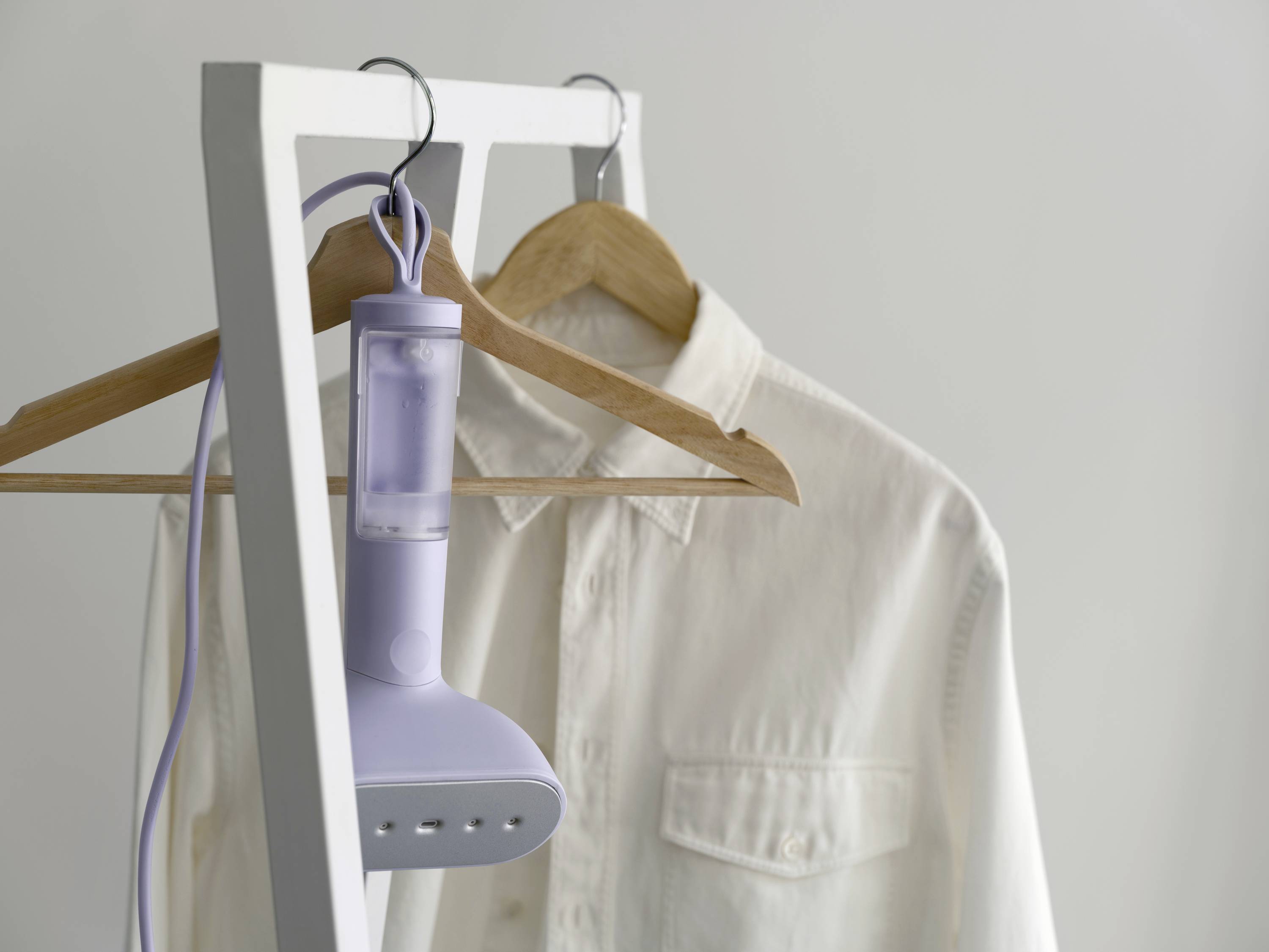 Ein weißes Hemd hängt auf einem Holzkleiderbügel an einem Kleiderständer. Ein Dampfbügeleisen ist am Bügel eingerollt.