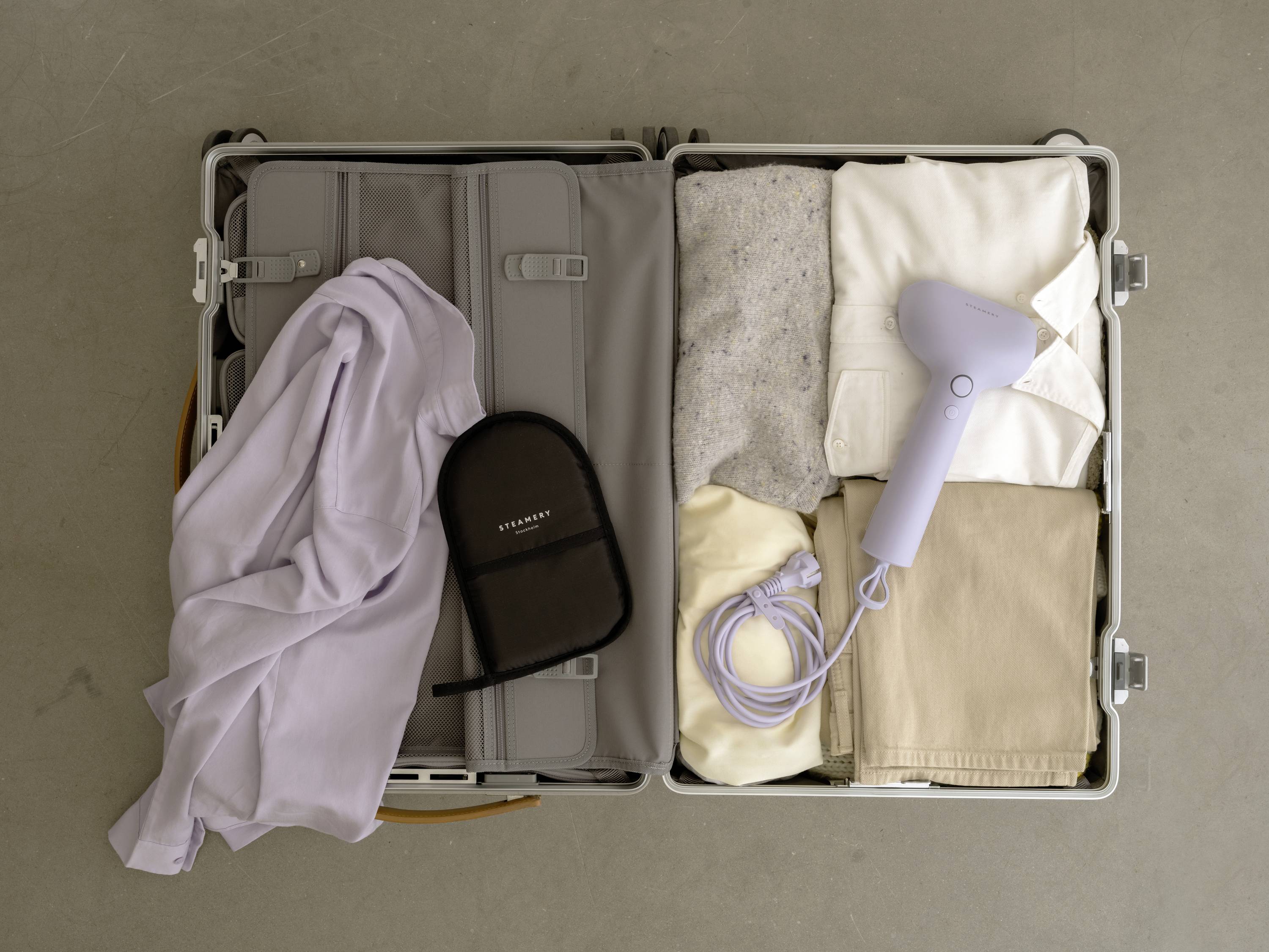 Ein offener Koffer zeigt ordentlich gepackte Kleidung und einen Föhn. Links liegt ein Hemd und ein Necessaire.