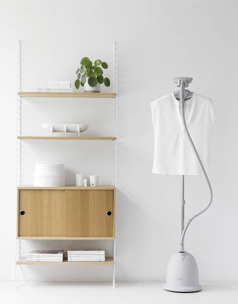 Ein Kleiderdampfer bearbeitet ein weißes Hemd. Daneben steht ein Regal mit minimalistischer Dekoration und Pflanzen.