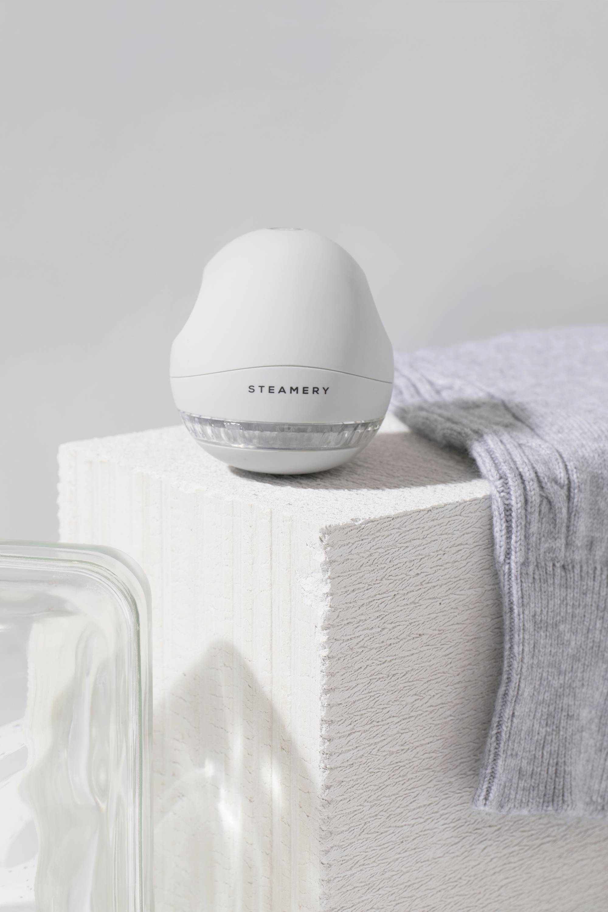 Ein weißer Textilentfussler von Steamery steht auf einem beigen Block neben einem grauen Kleidungsstück.