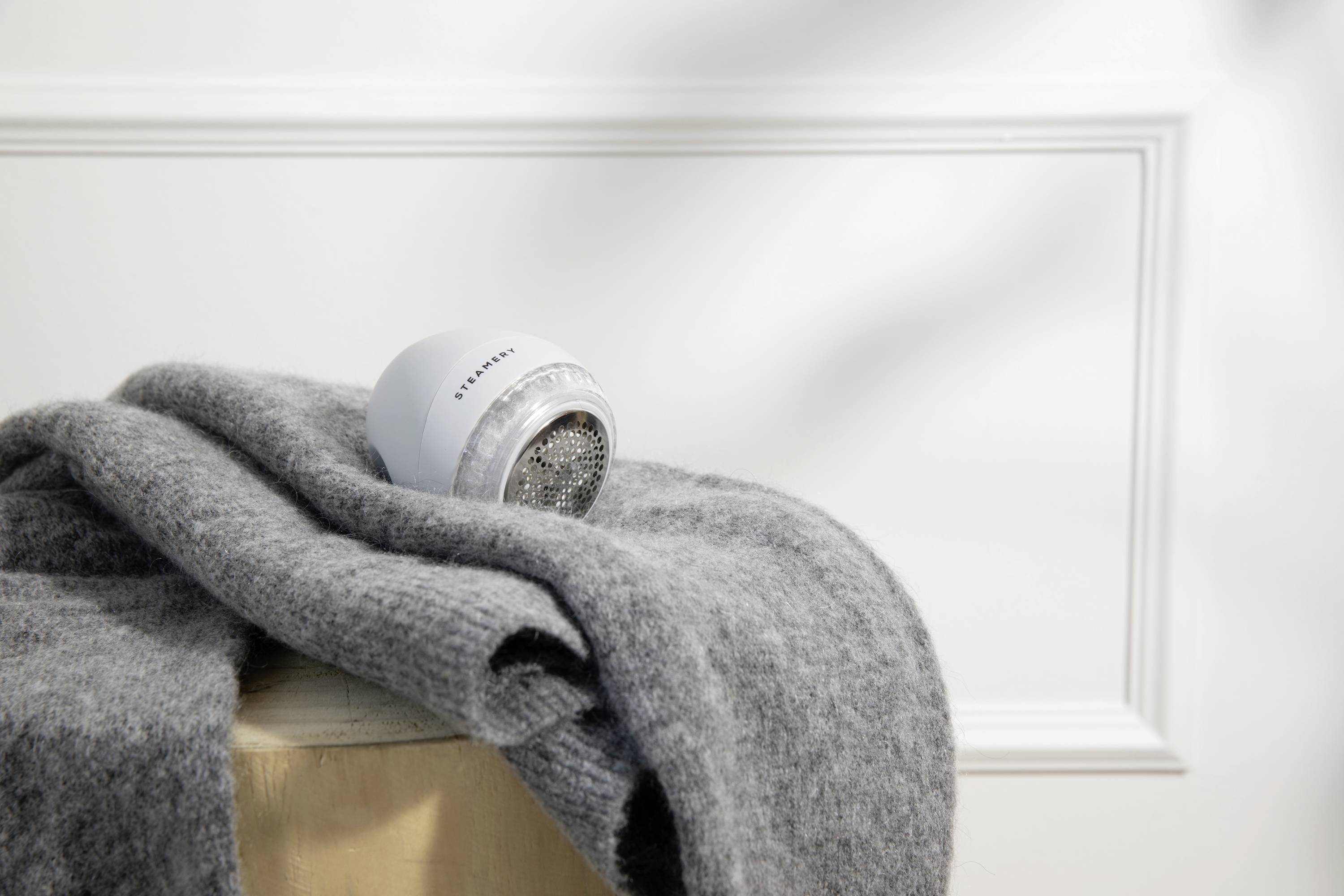 Ein grauer Pullover liegt auf einem Hocker, ein Fusselrasierer liegt oben drauf. Im Hintergrund ist eine weiße Wand zu sehen.