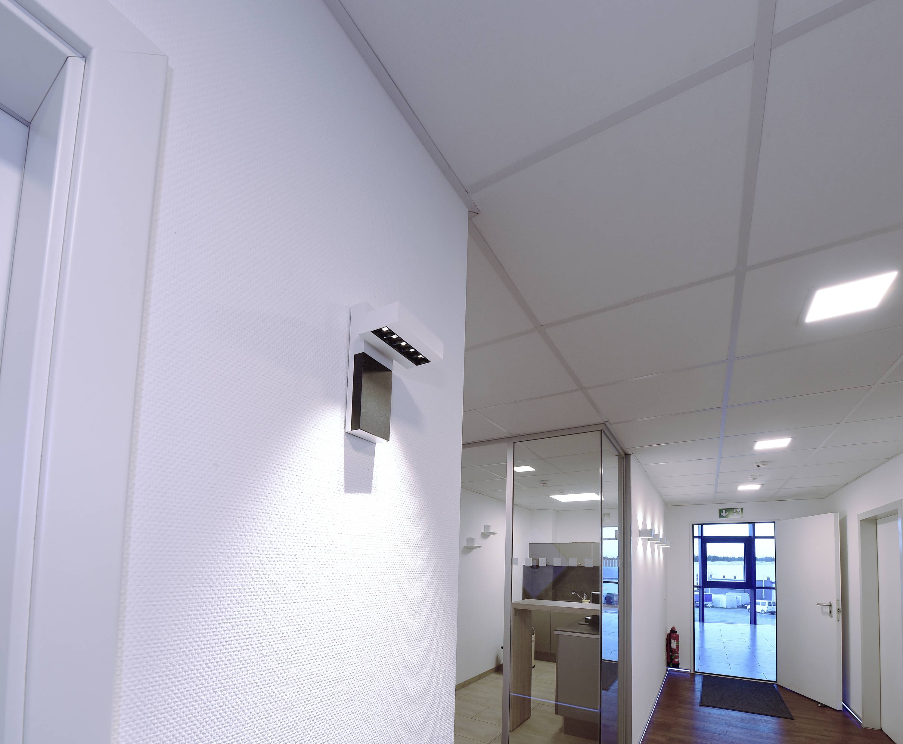 Gang in einem Bürogebäude mit beleuchteter Wand und Fenster am Ende. Lampen und Türen entlang des Weges, ein Feuerlöscher rechts.