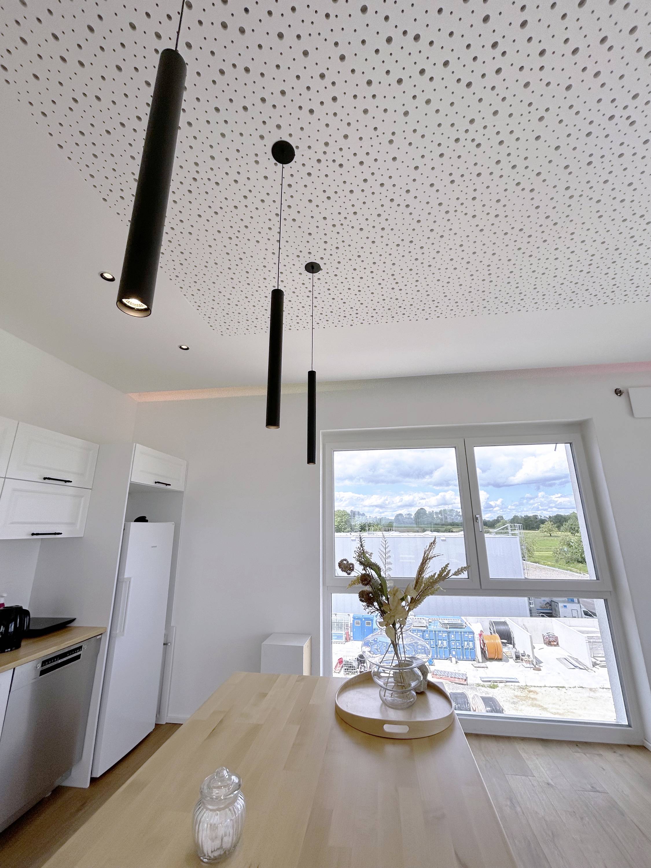 Moderne Küche mit hellem Holztisch, weißen Schränken und drei schwarzen Hängelampen. Durch das Fenster ist eine Baustelle sichtbar.