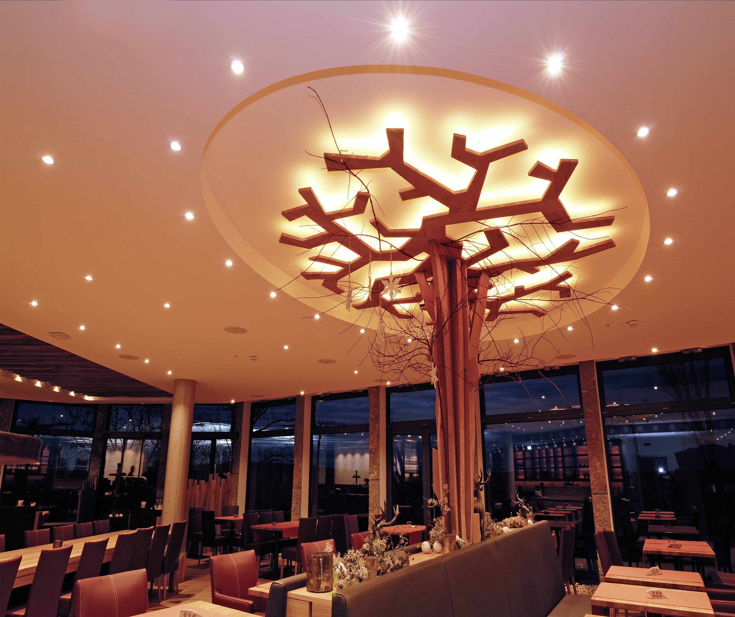 Beleuchtetes Restaurant mit dekorativem, baumförmigem Leuchter an der Decke, dunkles Interieur und große Fenster im Hintergrund.