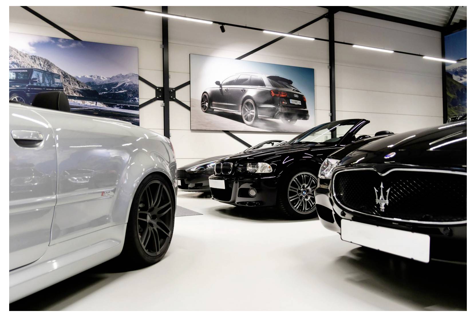 Drei luxuriöse Sportwagen in einem Showroom ausgestellt, darunter ein schwarzer Maserati im Vordergrund und gerahmte Autofotos an der Wand.