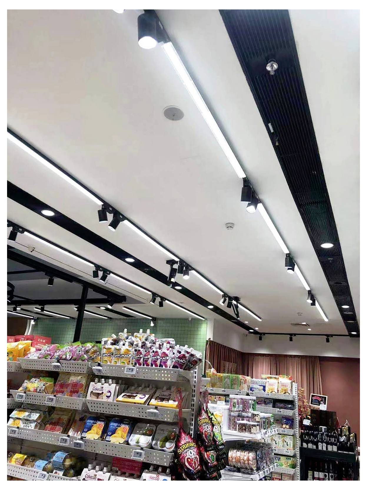 Innenansicht eines Gangs in einem Supermarkt mit Regalen voller Snacks, Getränke und Haushaltswaren unter hellen Deckenlampen.