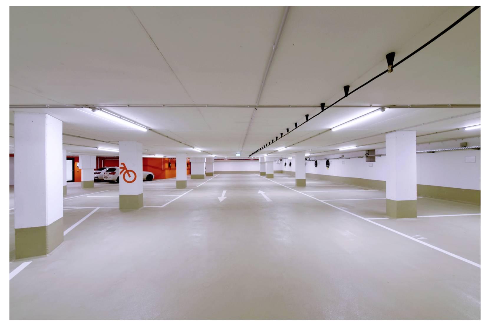 Leere unterirdische Tiefgarage mit weißen Säulen, markierten Stellplätzen und einem geparkten Auto in der Ferne.