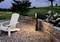 'Weißer Gartenstuhl auf einem Kiesboden neben einer beleuchteten Gartenlampe, umgeben von Blumenbeeten und Steinmauer im Abendlicht.'