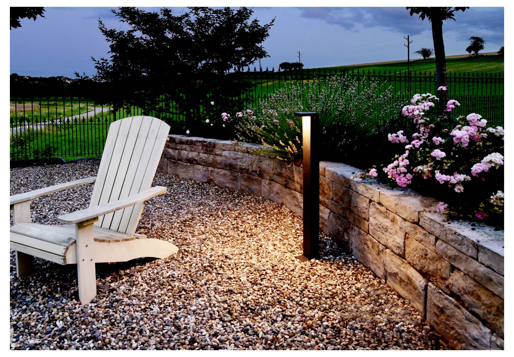 Weißer Holzstuhl neben einem beleuchteten Wegpfosten in einem angelegten Garten mit Natursteinmauer, Blumen und eingezäuntem Grashang in der Dämmerung.