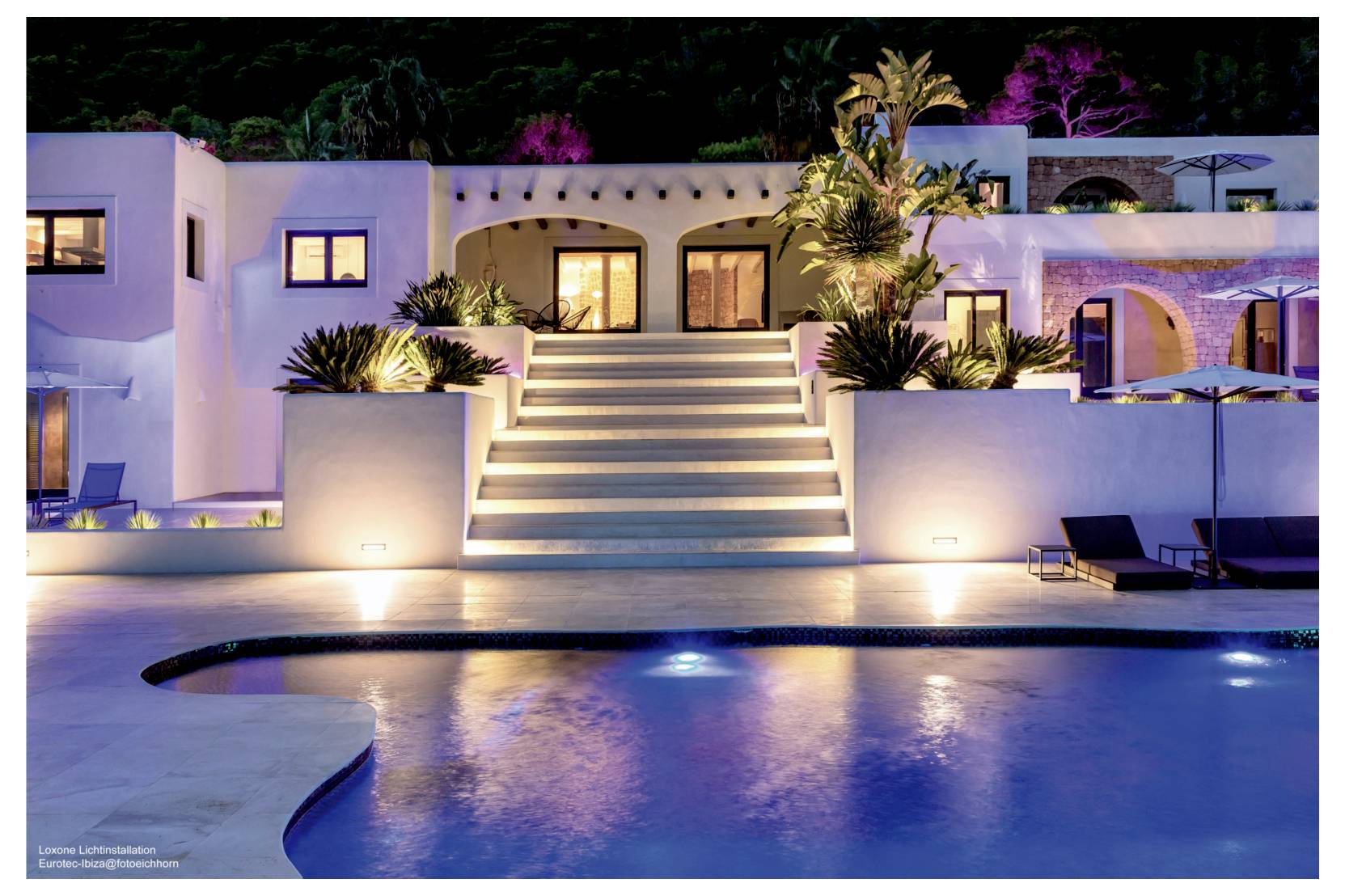 Stilvolles Ferienhaus im Stil eines Resorts bei Nacht mit beleuchteter zentraler Treppe, Palmen und einem Swimmingpool davor.