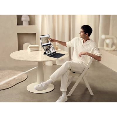 Ein Mann sitzt an einem runden Tisch und tippt auf einen Laptop, der Videoanrufe und Diagramme anzeigt. Die Umgebung ist minimalistisch gestaltet.