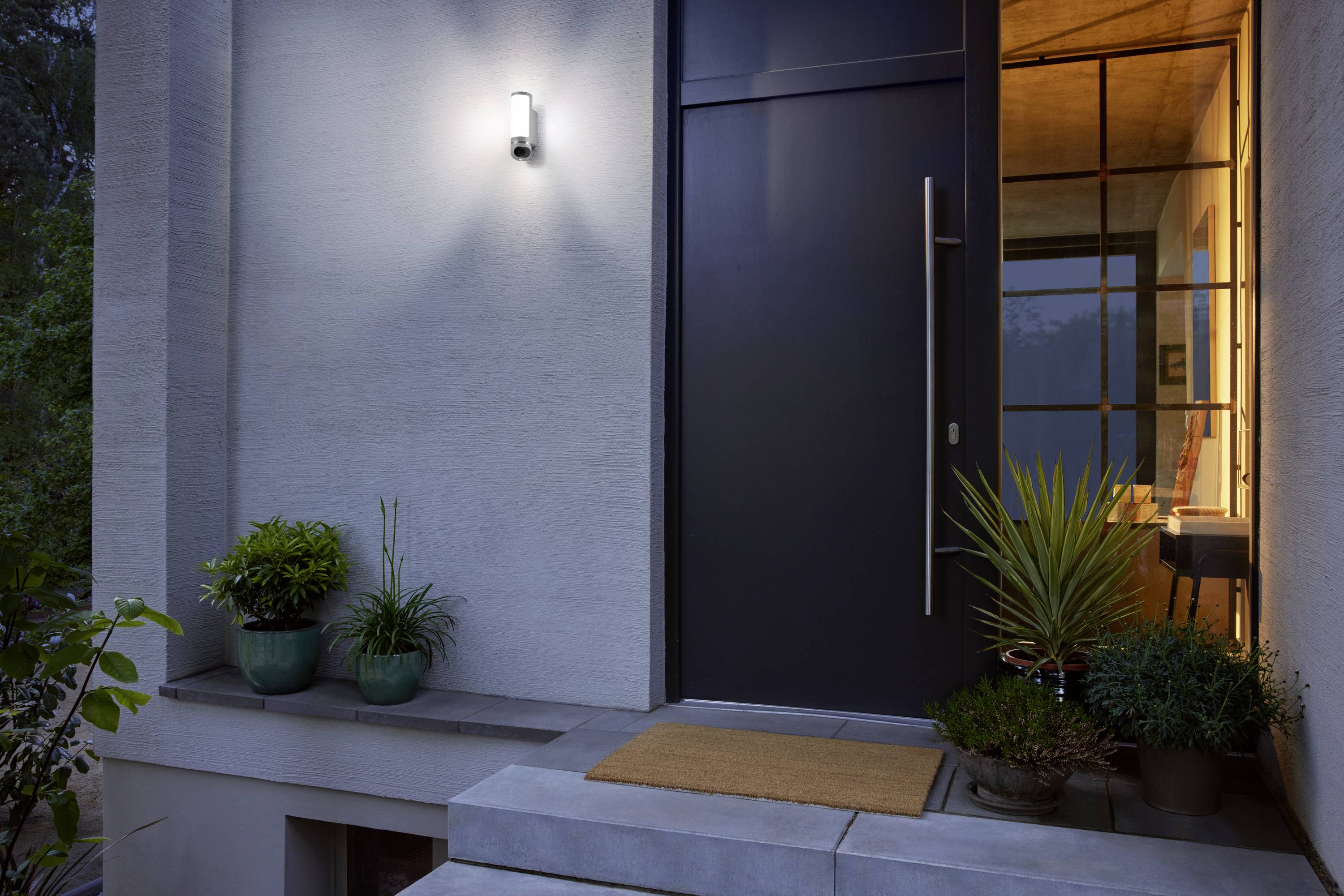 Ein modern gestalteter Hauseingang bei Dämmerung, beleuchtet von einer Wandlampe, mit Topfpflanzen und einer braunen Fußmatte.