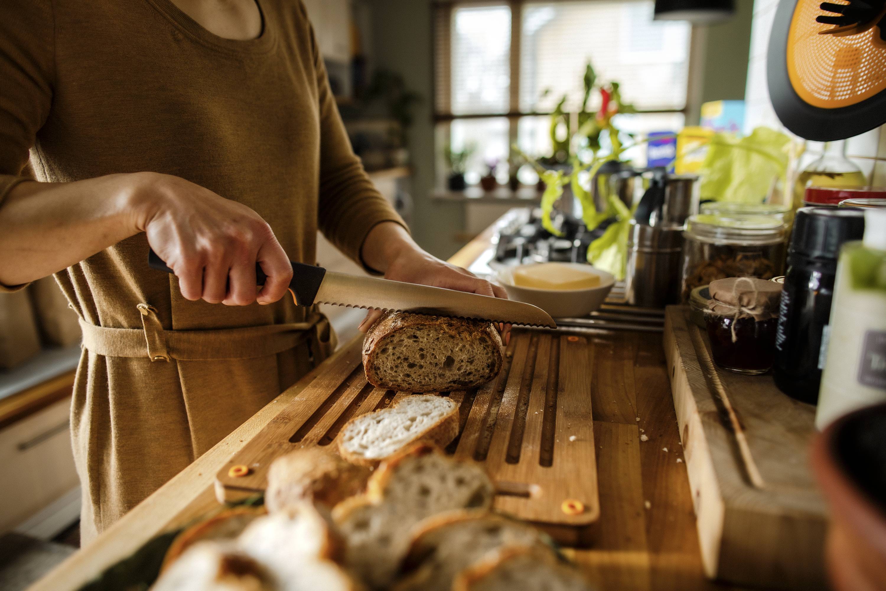 Person schneidet Brot in einer Küche. Auf dem Tisch liegen bereits mehrere Brotscheiben. Im Hintergrund sind Küchengeräte sichtbar.