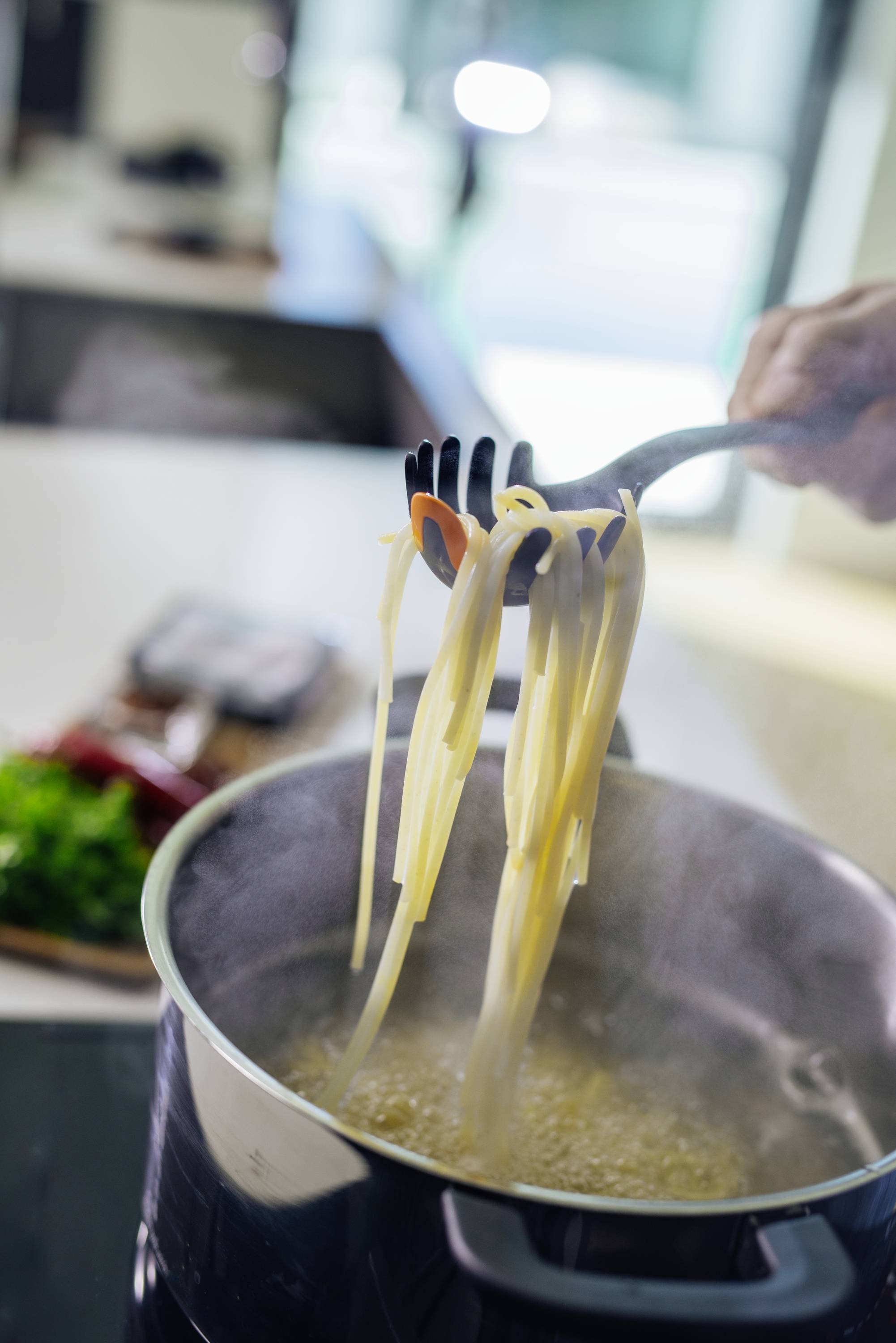 Eine Hand hebt mit einer Gabel Spaghetti aus einem kochenden Topf in einer modernen Küche. Im Hintergrund sind Zutaten sichtbar.