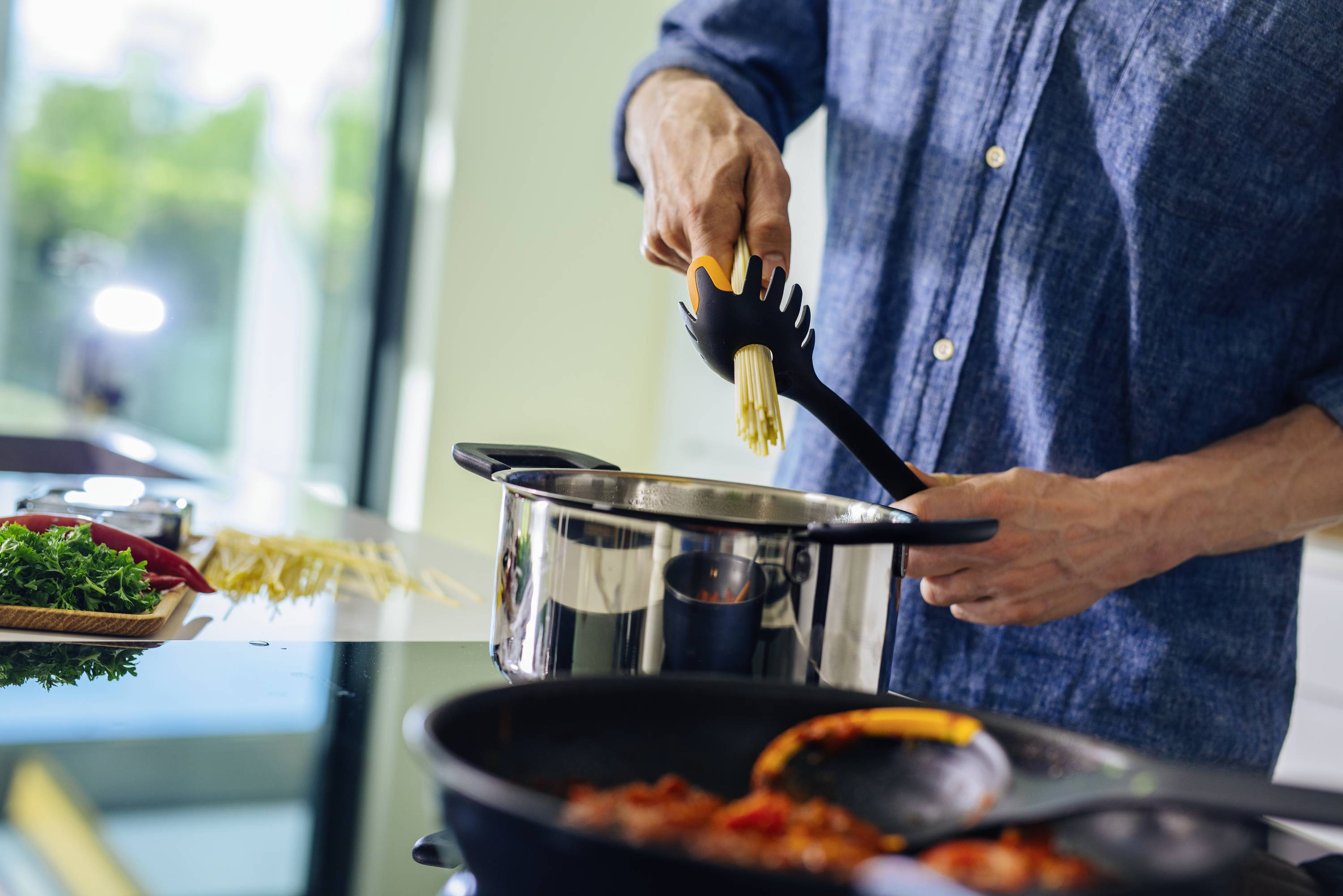 Person kocht in moderner Küche und gibt Nudeln in einen Topf auf einem Herd. Im Vordergrund sind Gewürze und eine Pfanne zu sehen.