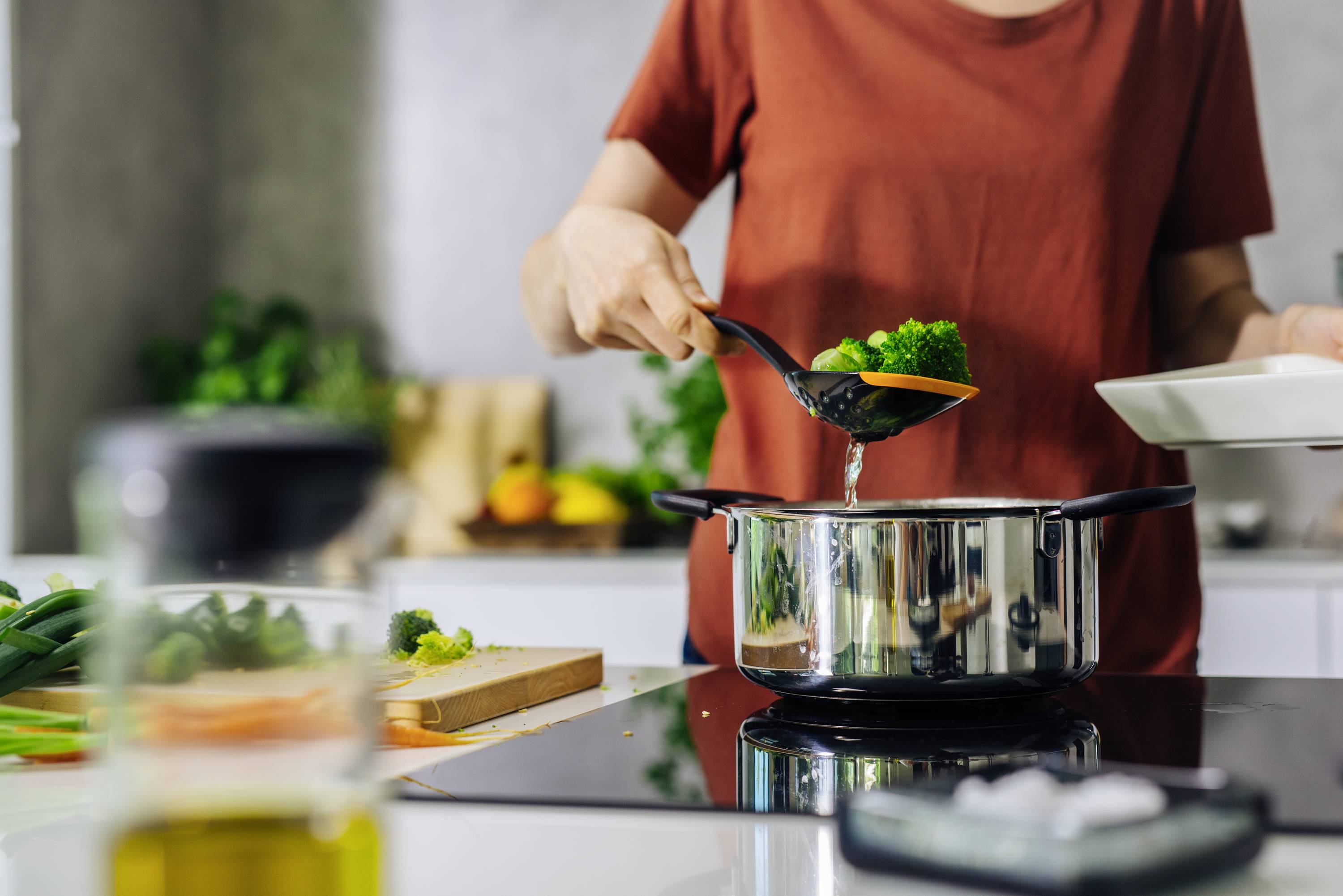 Person kocht in einer modernen Küche, gibt Gemüse in einen Kochtopf auf dem Herd. Frisches Gemüse und Olivenöl im Hintergrund.