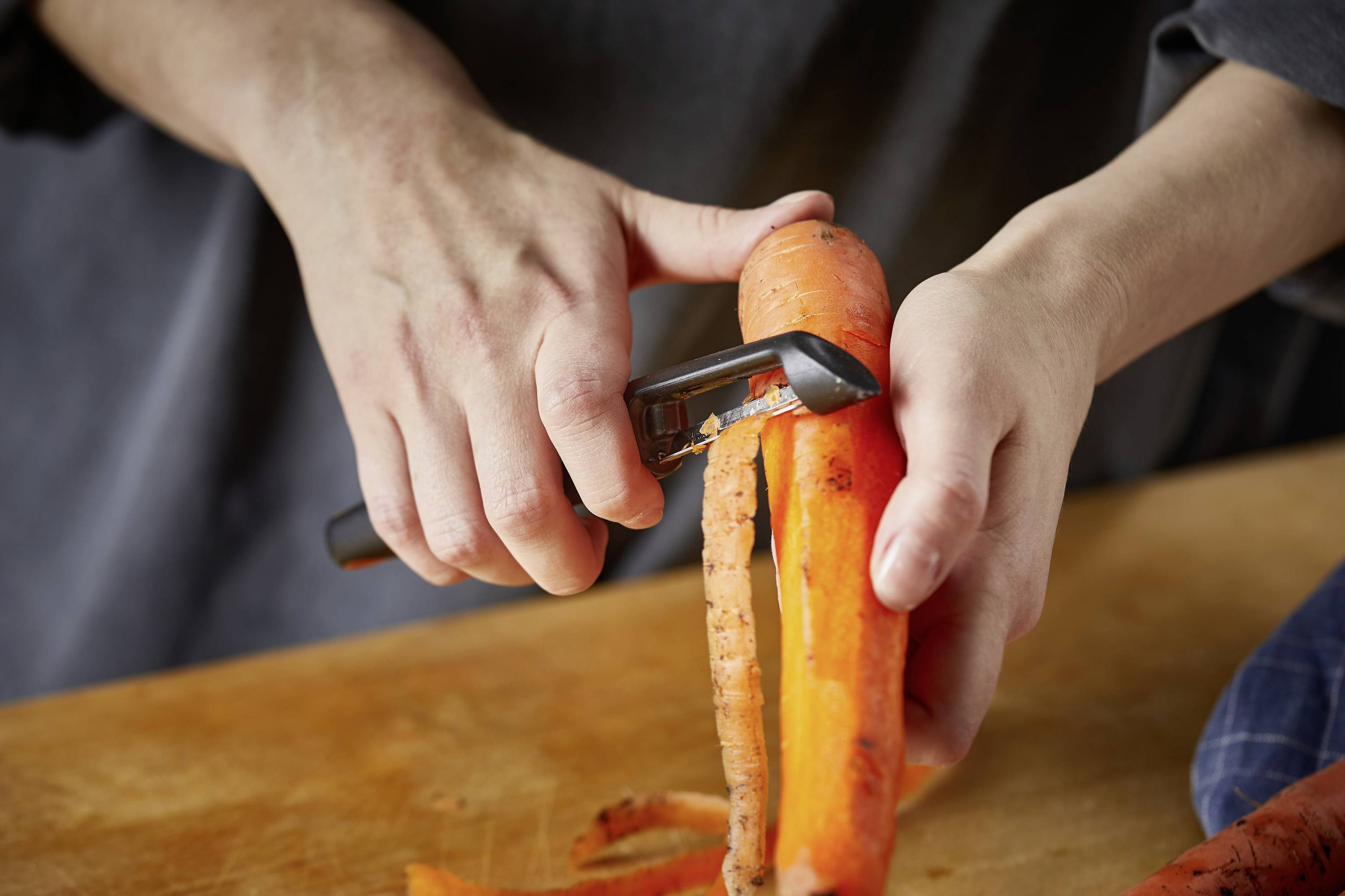 Person schält eine Karotte mit einem Sparschäler auf einem Holzbrett. Hände und Werkzeug sind im Fokus, Küchentätigkeit erkennbar.