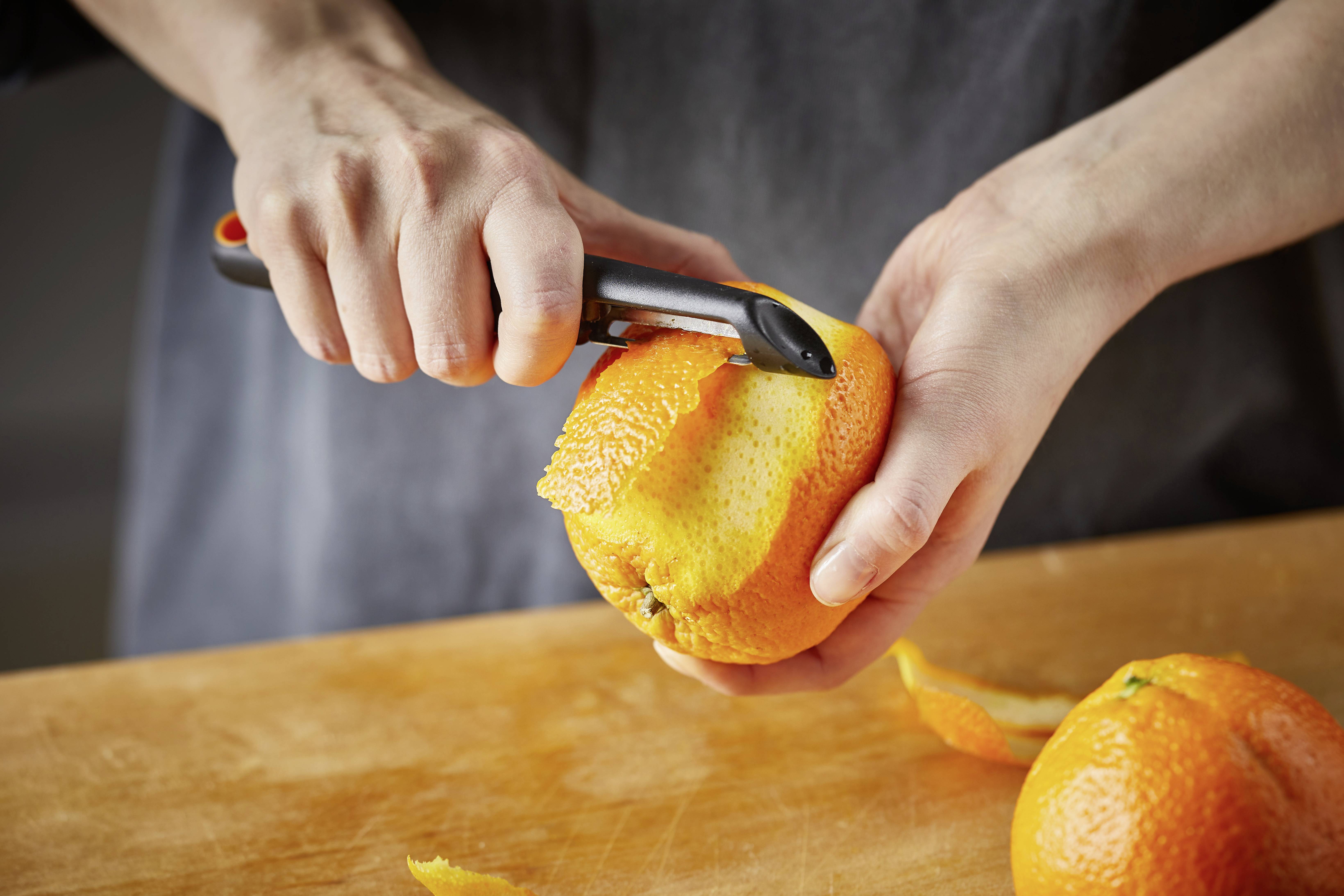 Eine Person schält eine Orange mit einem Schäler auf einem Holzbrett.