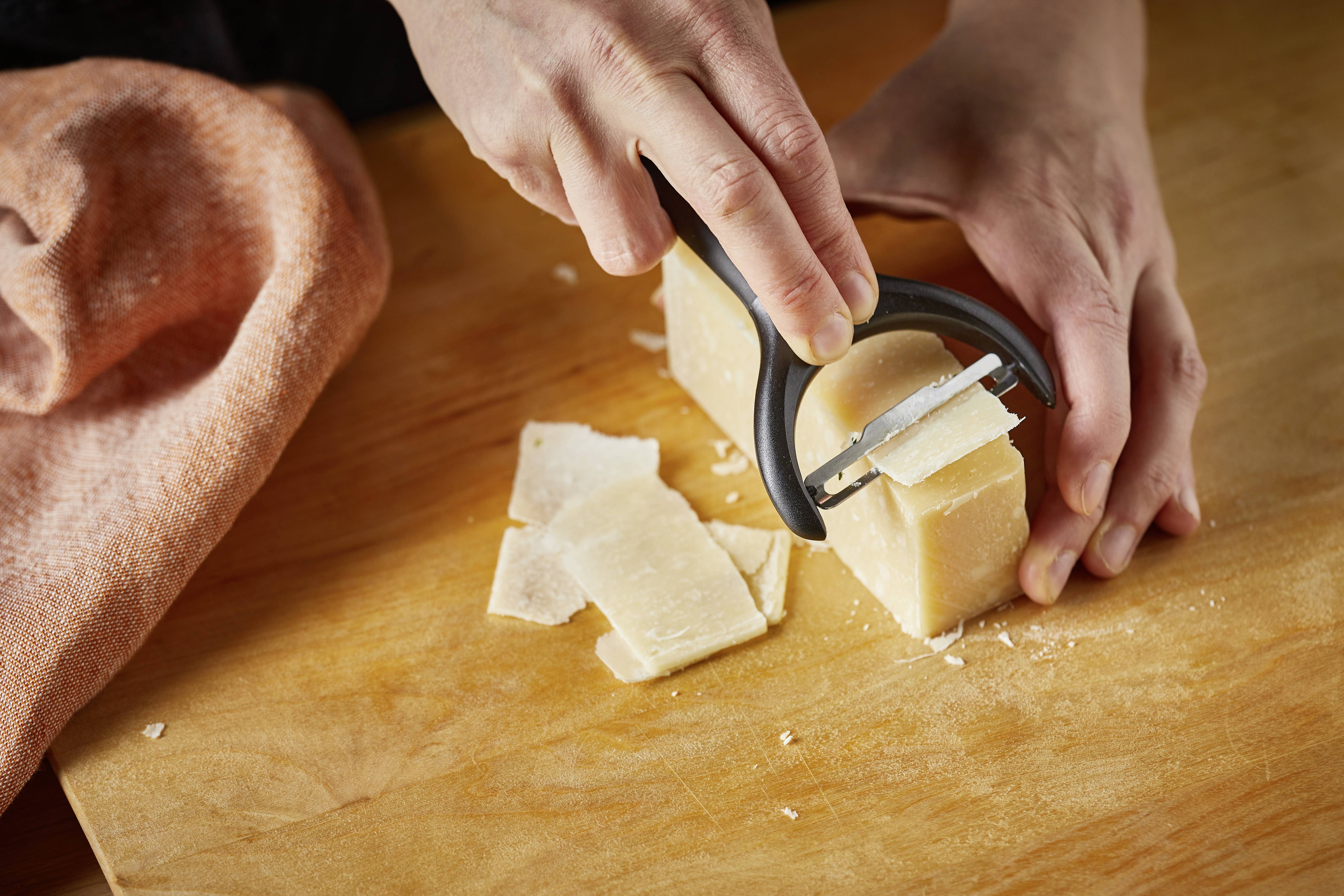 Eine Person reibt Parmesan mit einem schwarzen Handhobler, während Käseflocken auf einem hölzernen Schneidebrett landen.