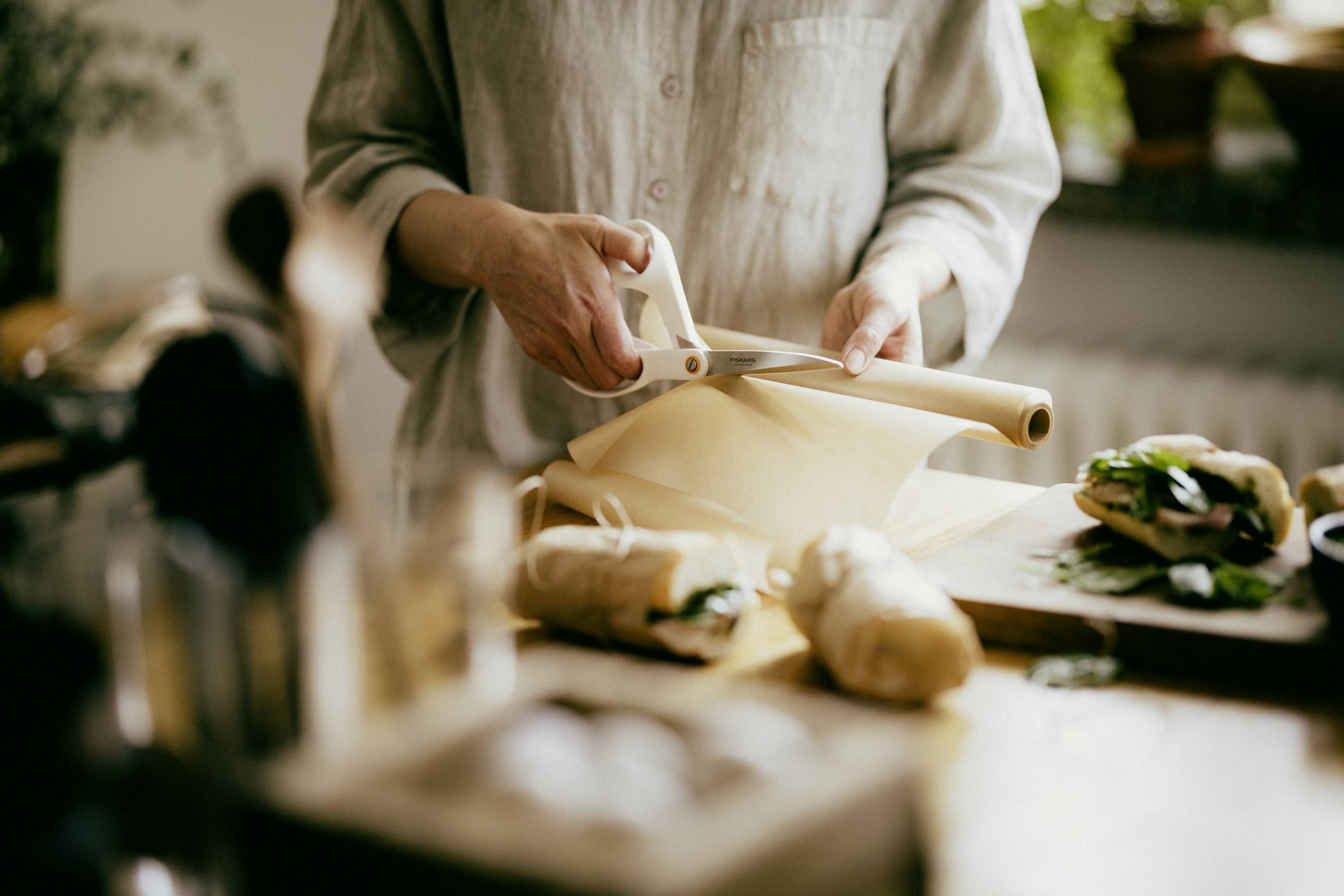 Person schneidet Backpapier in der Küche. Nebenbei liegen zwei eingewickelte Sandwiches. Warmes Licht und Pflanzen im Hintergrund.