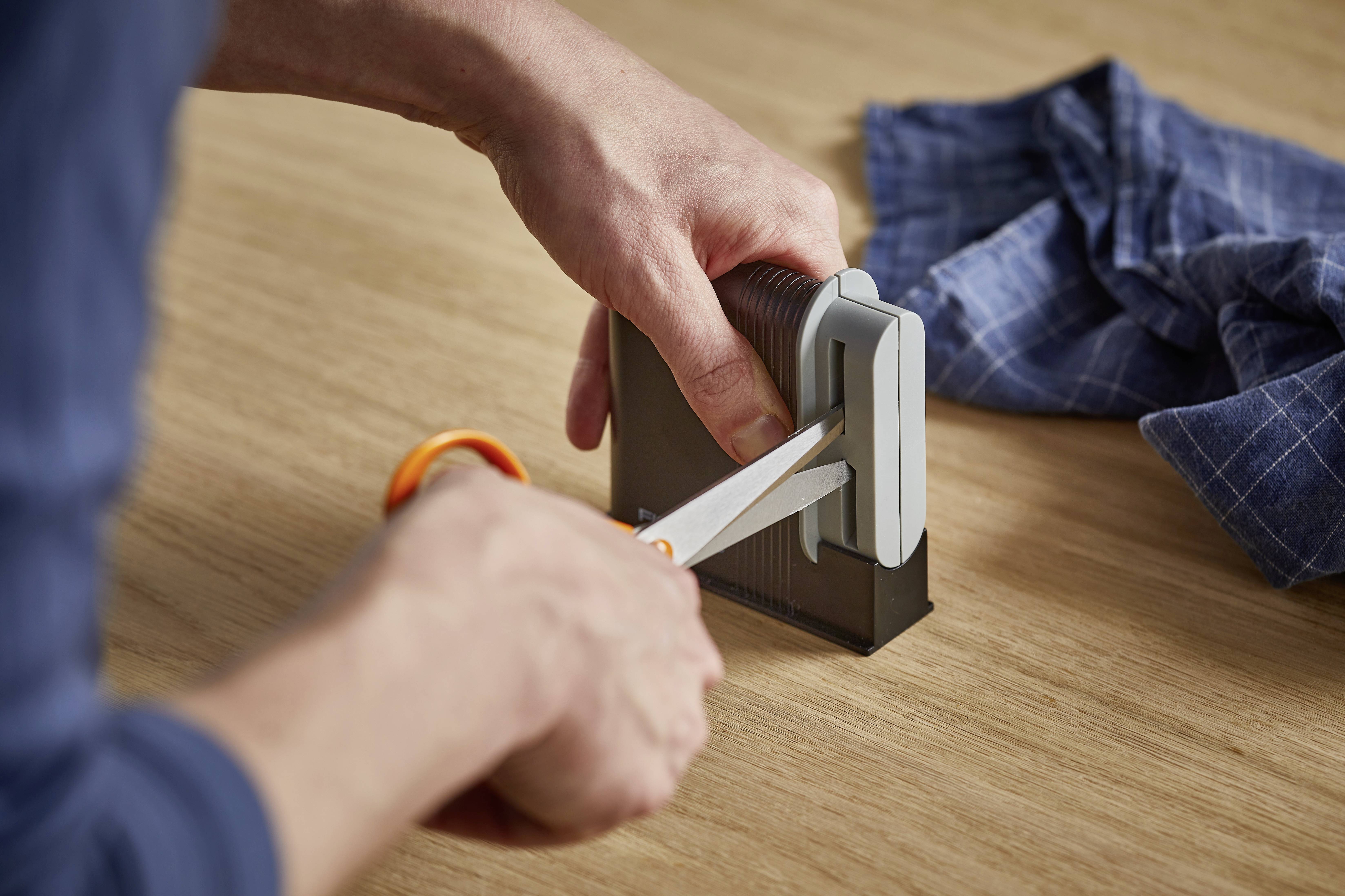 Eine Person schärft eine Schere mit einem Schärfgerät auf einem Holztisch. Ein blau kariertes Tuch liegt daneben.