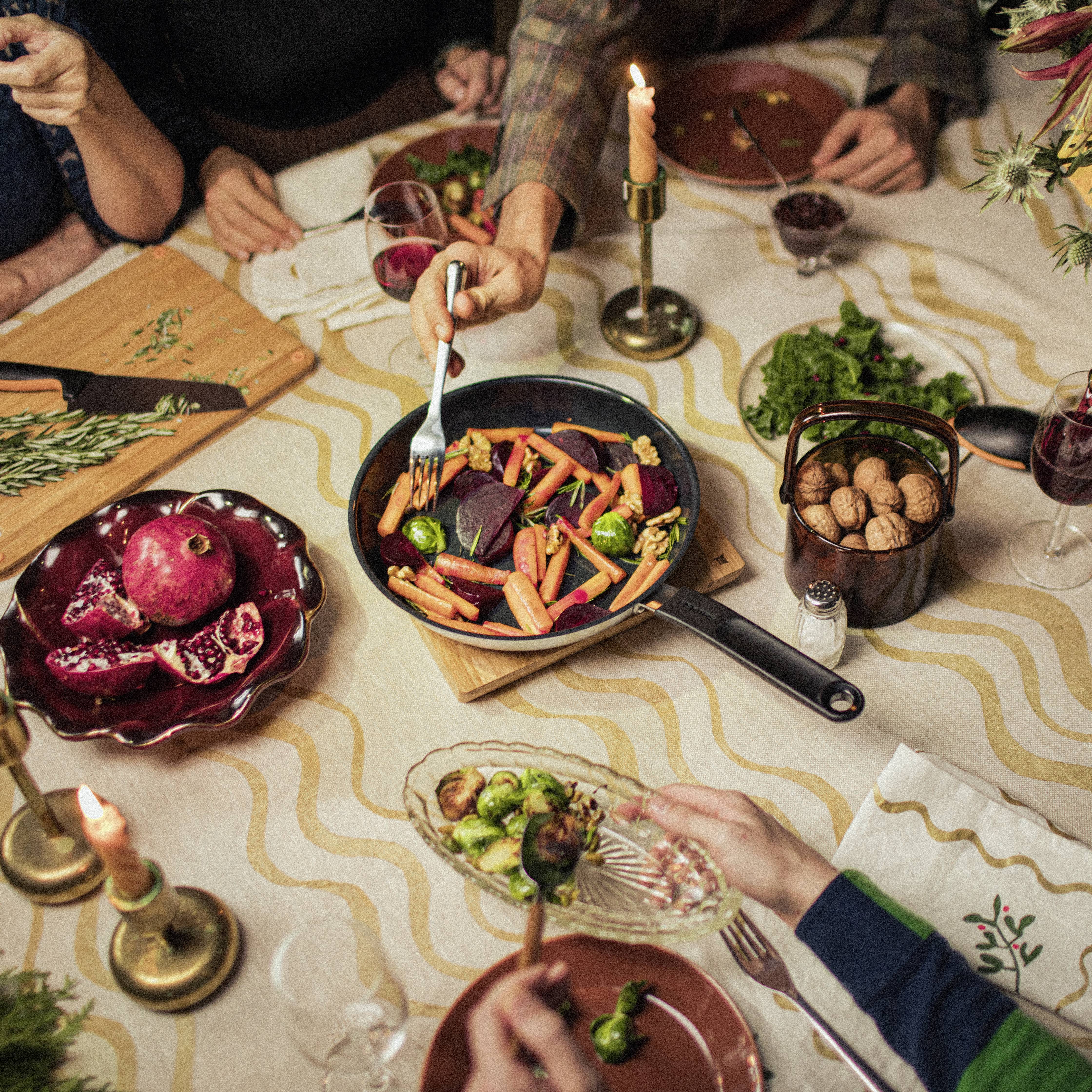 Mehrere Personen sitzen an einem Tisch beim Abendessen. Im Zentrum steht eine Pfanne mit buntem Gemüse. Kerzen und Gewürze sind dekorativ arrangiert.